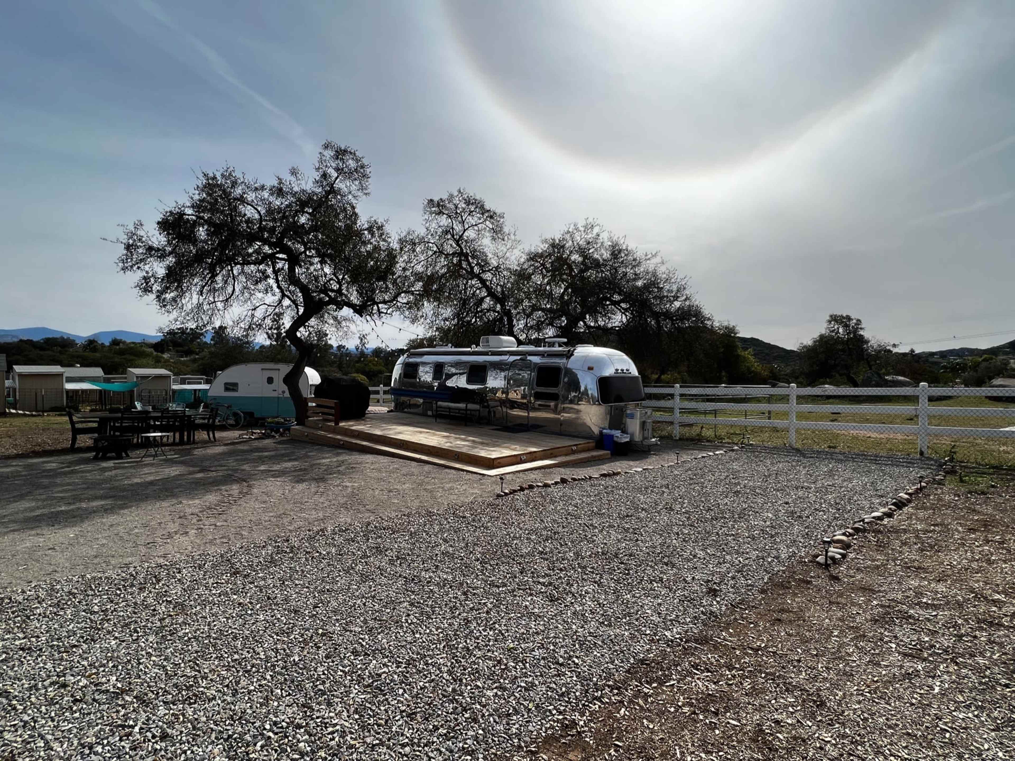 An Airstream trailer is parked on a gravel area under a large tree, with a wooden deck and a fenced field in the background.