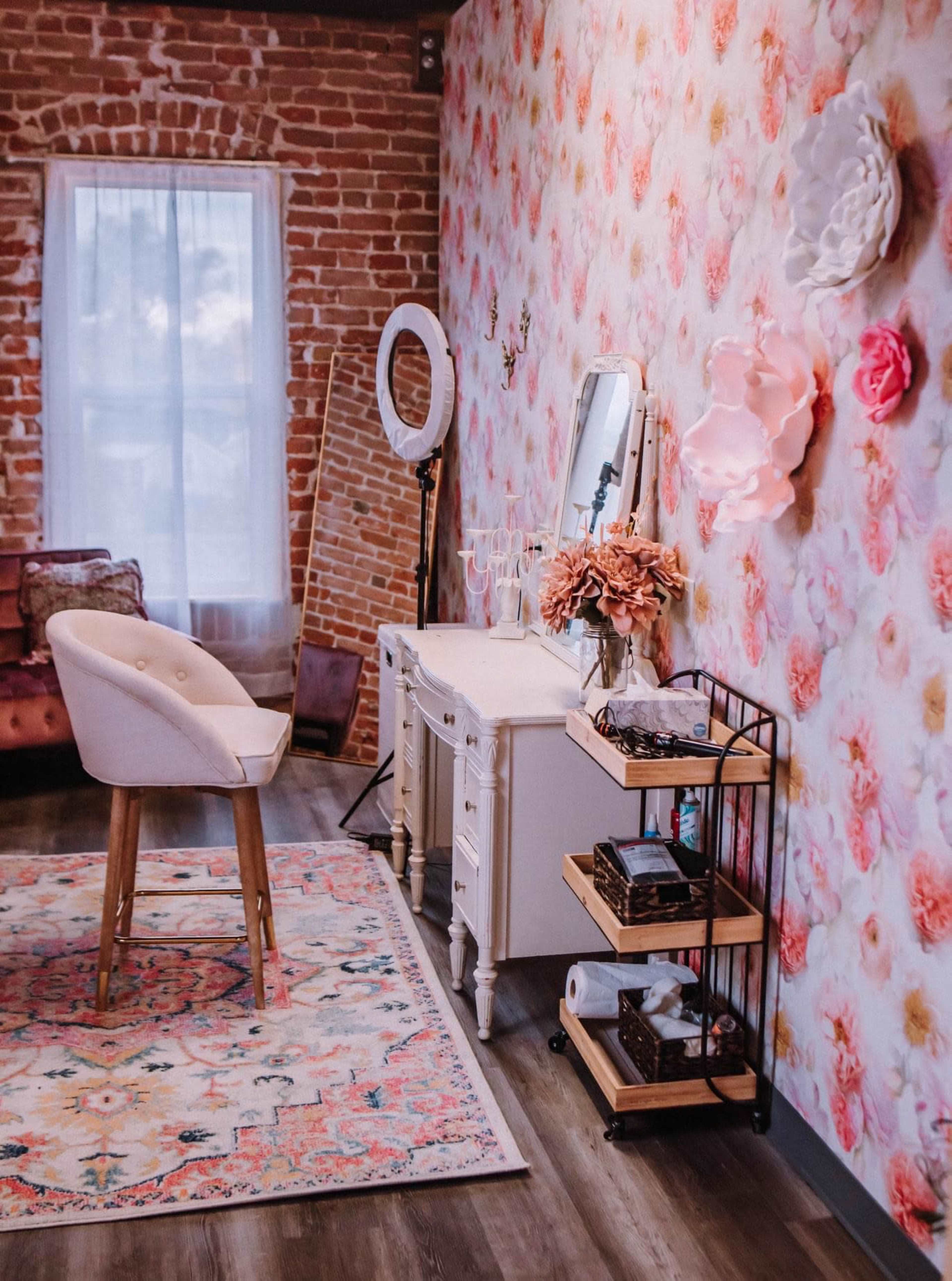 A stylized room with a floral wallpaper, a white vanity table with a mirror, a chair, and a cart displaying various items, all set against a backdrop of exposed brick and a colorful area rug.