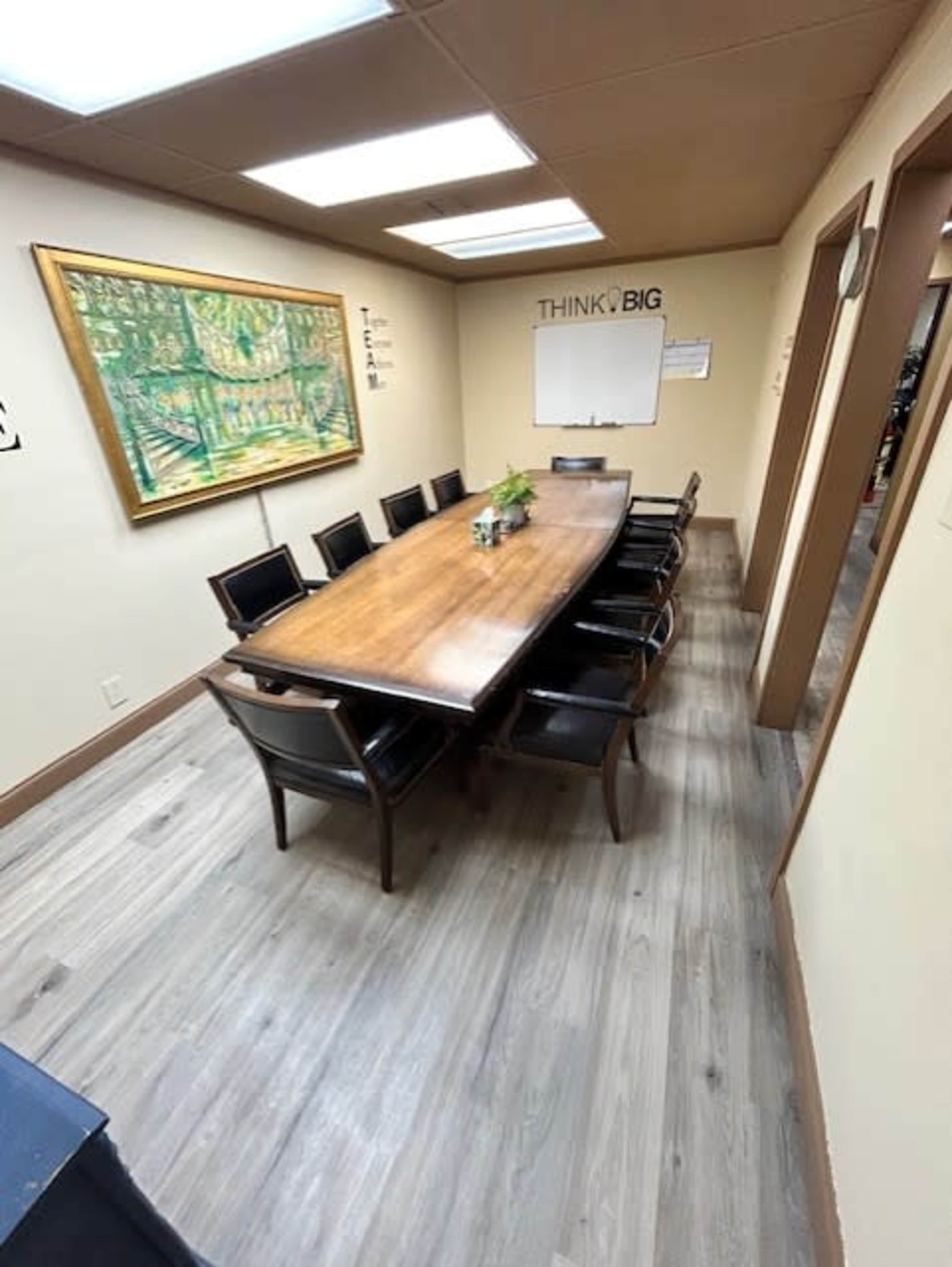 A conference room with a long wooden table surrounded by eight black chairs, featuring a large painting on the wall and a whiteboard.