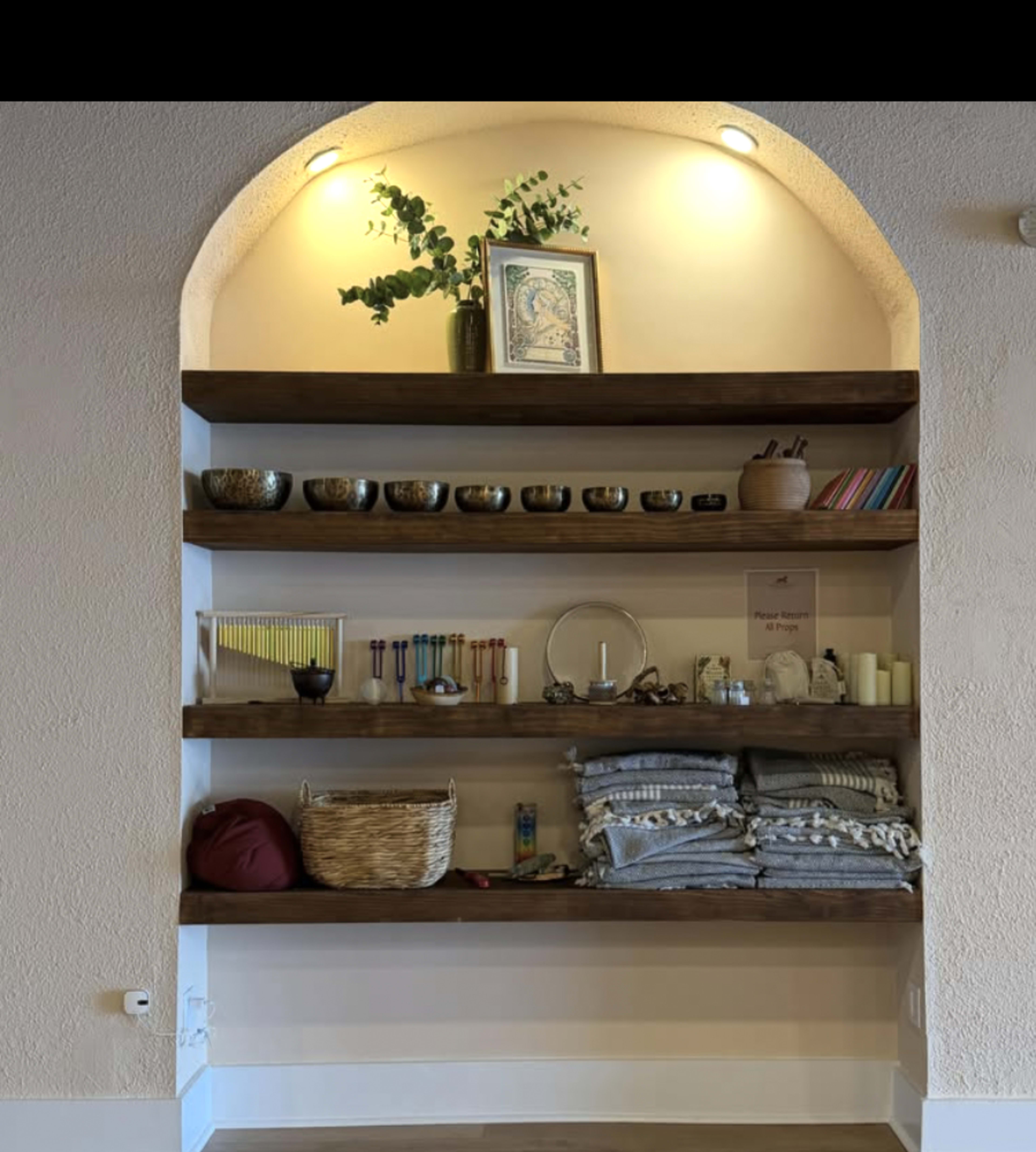 The image shows a well-organized shelf with various items including decorative bowls, musical instruments, books, candles, and neatly folded blankets, set within an alcove illuminated by lights.