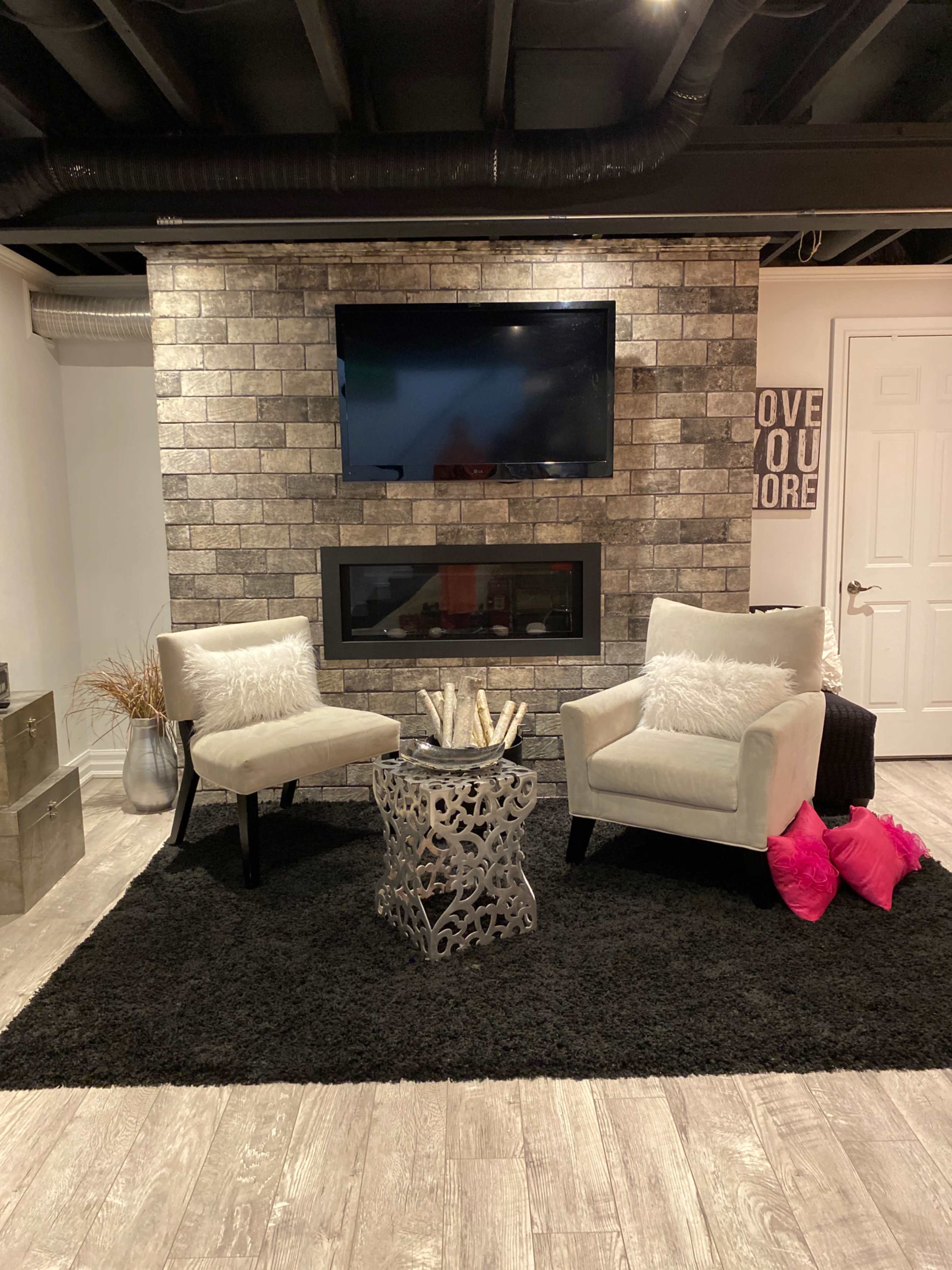 A modern living room with two white armchairs facing a wall-mounted television and an electric fireplace, accompanied by a decorative table and pillows on a black rug.