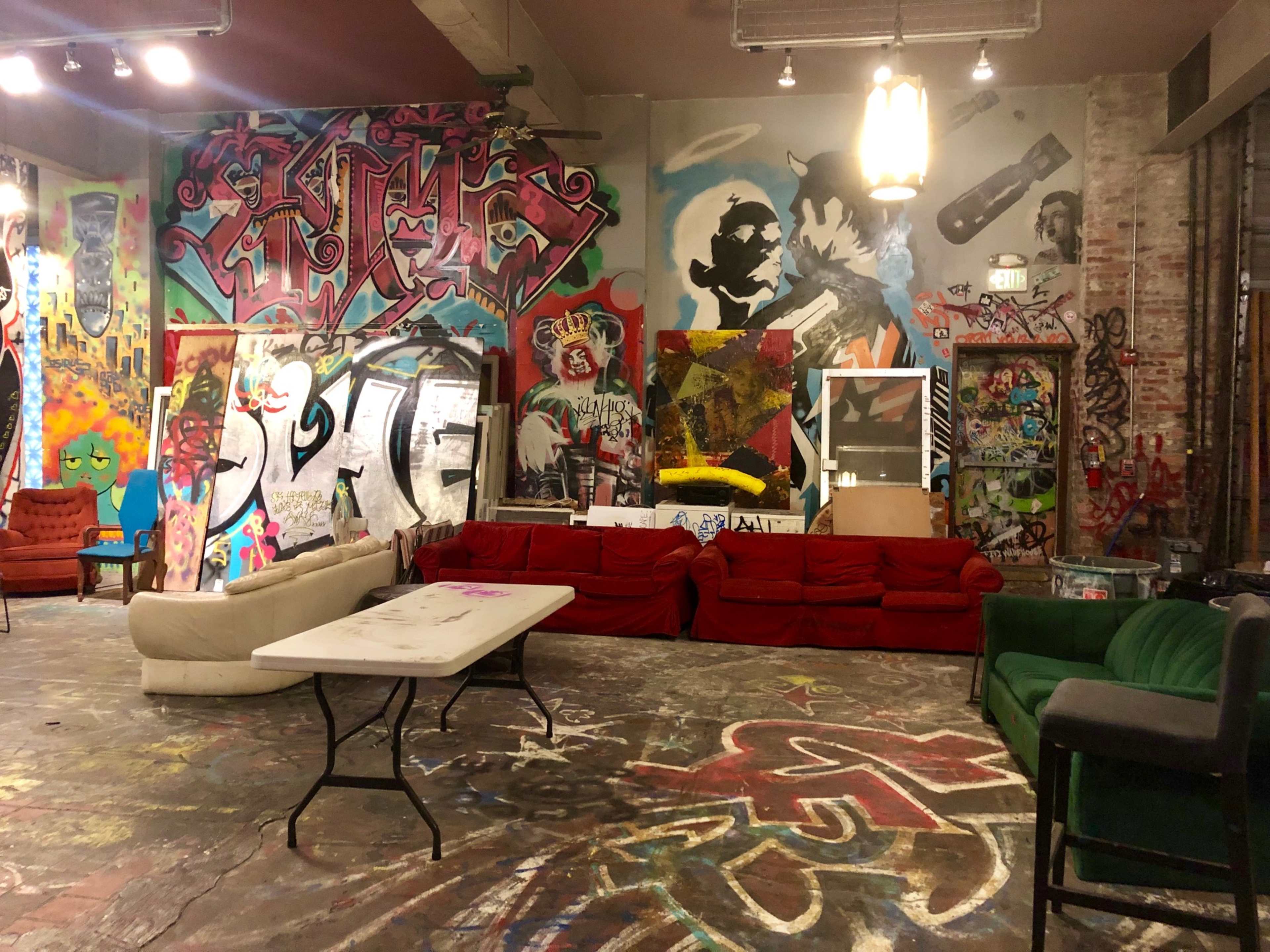 An interior space featuring colorful graffiti-covered walls, several couches, and a table in the center of a room with a concrete floor.