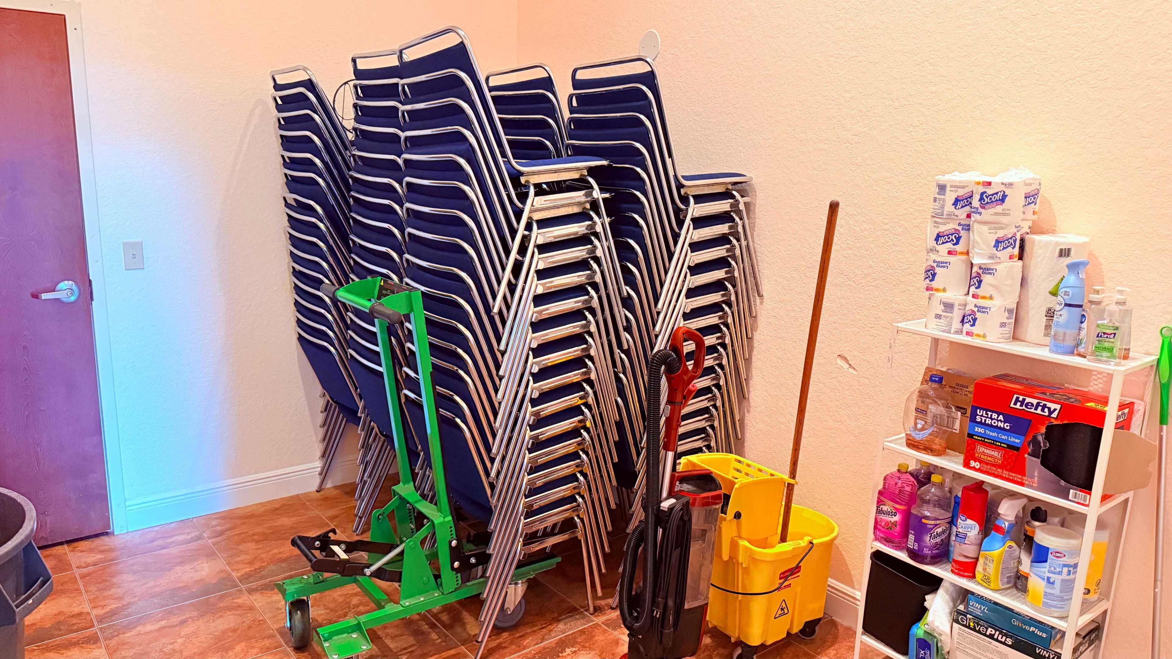 The image shows a stack of blue folding chairs next to cleaning supplies and a yellow mop bucket in a room with beige walls and tiled flooring.