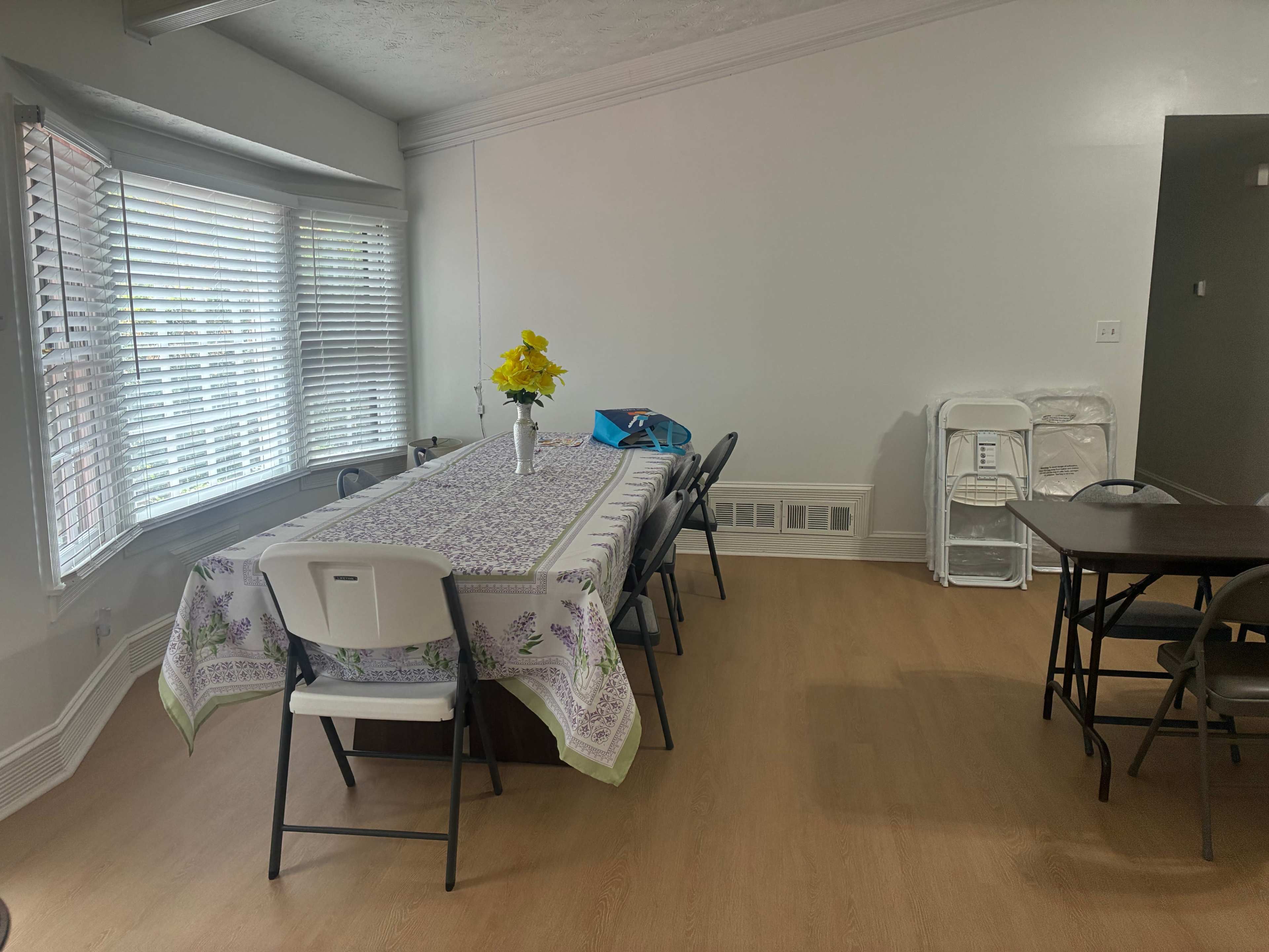 A spacious room features a long table with a floral tablecloth, surrounded by folding chairs, and a window with blinds allowing natural light to enter.