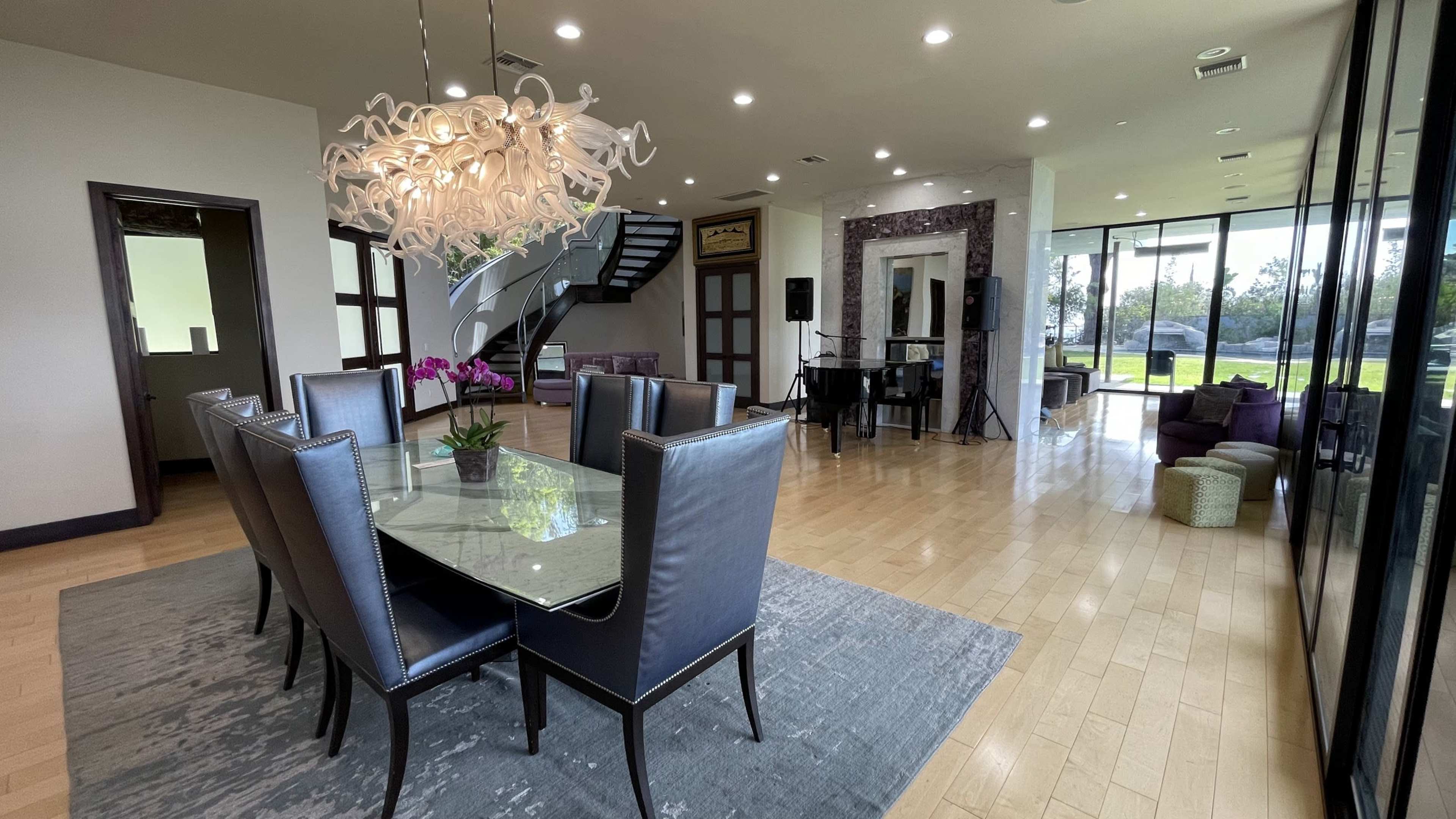 A modern dining area features a glass table surrounded by black chairs, with a staircase in the background and large windows overlooking a landscaped area.