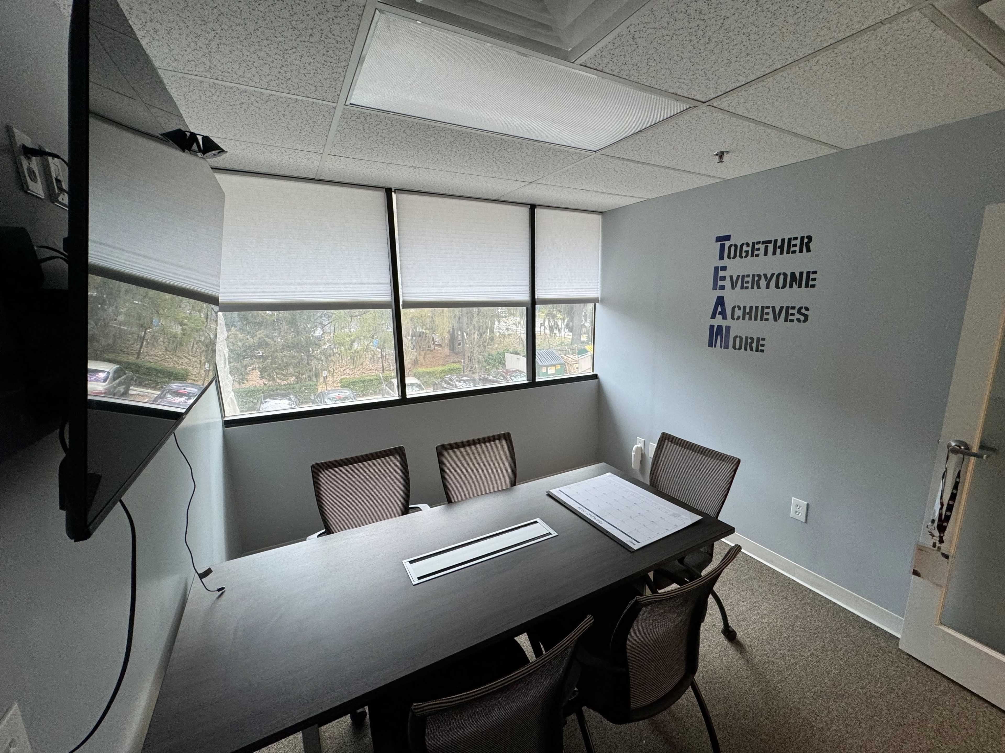 A small conference room with a long table and four chairs, featuring large windows that allow natural light and a wall with the word "TEAM" creatively displayed alongside the phrase "Together Everyone Achieves More."
