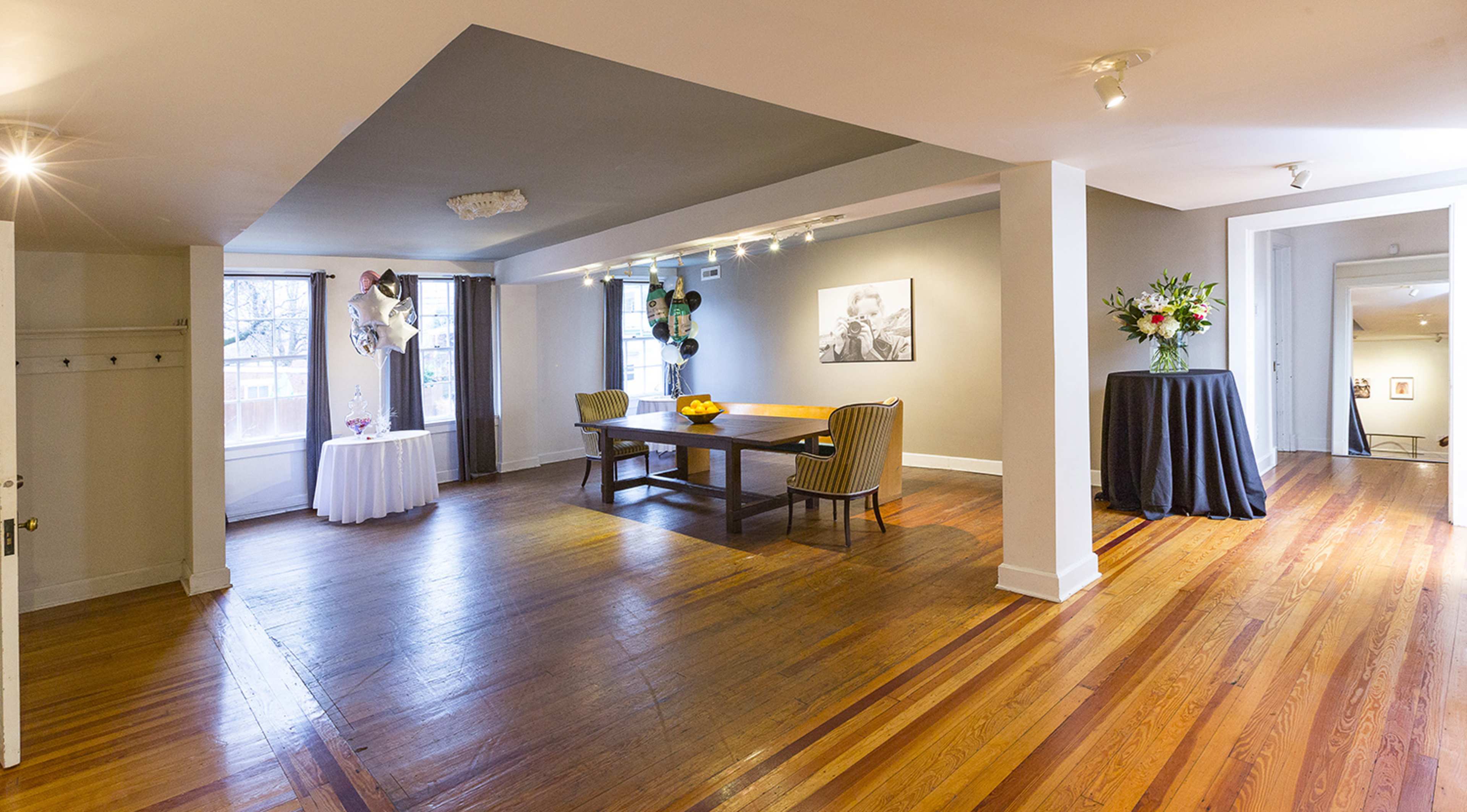 A spacious room with wooden floors, featuring a large table, chairs, and floral arrangements, along with multiple windows letting in natural light.