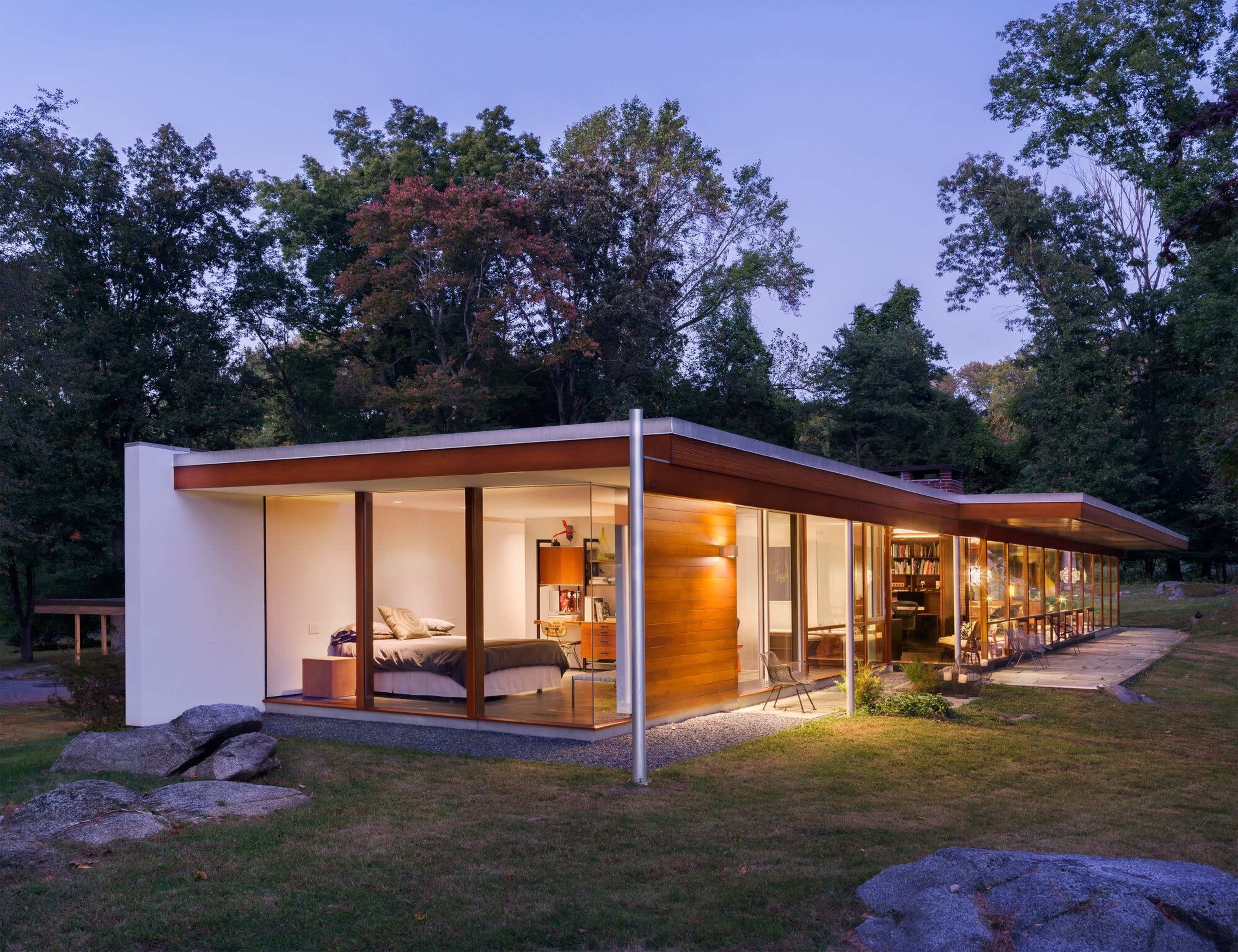 A modern house with large glass windows and a flat roof is set amidst trees, showcasing a well-lit interior and a landscaped yard.