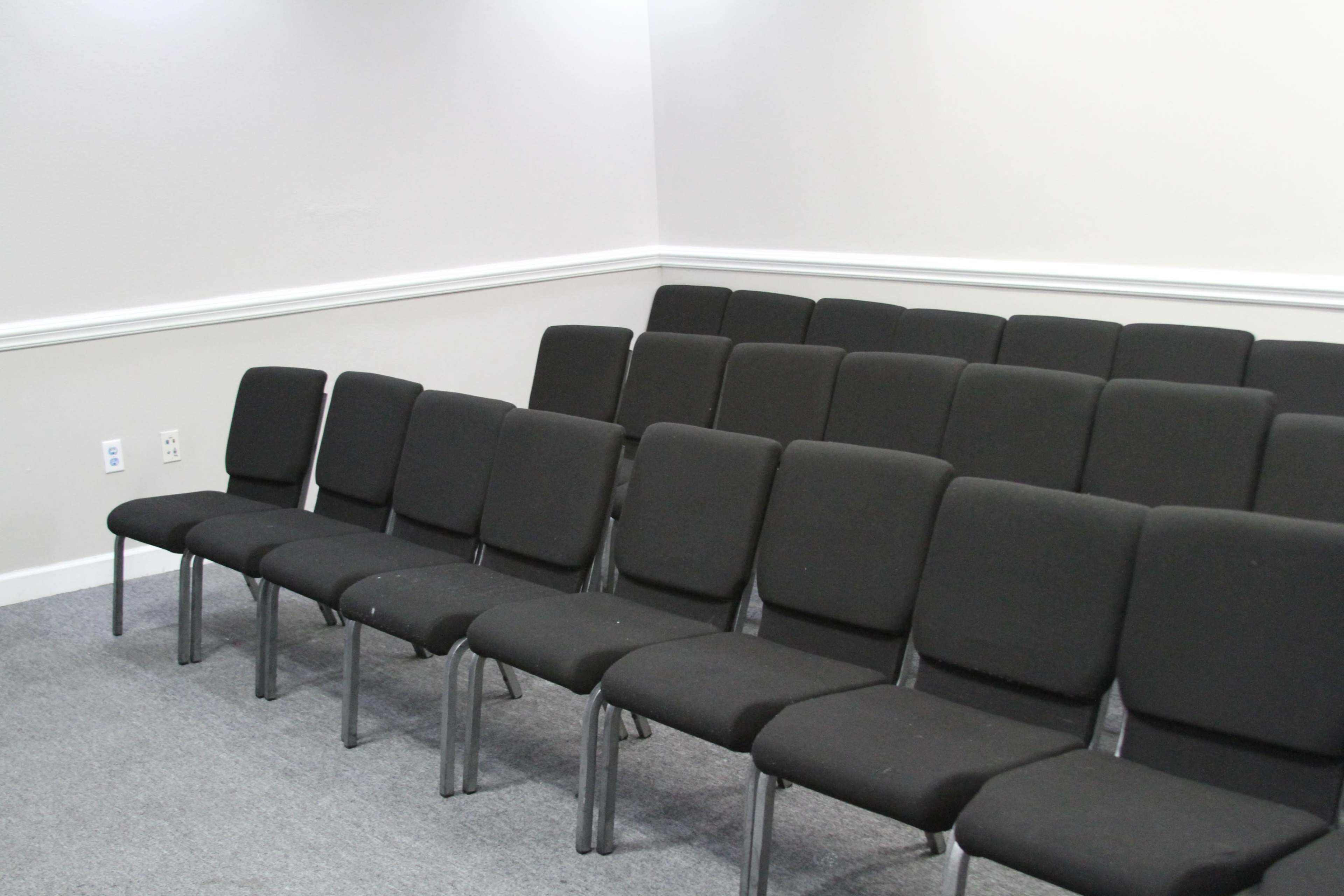 A row of black, upholstered chairs arranged in a sparsely decorated room with light-colored walls and a gray carpet.