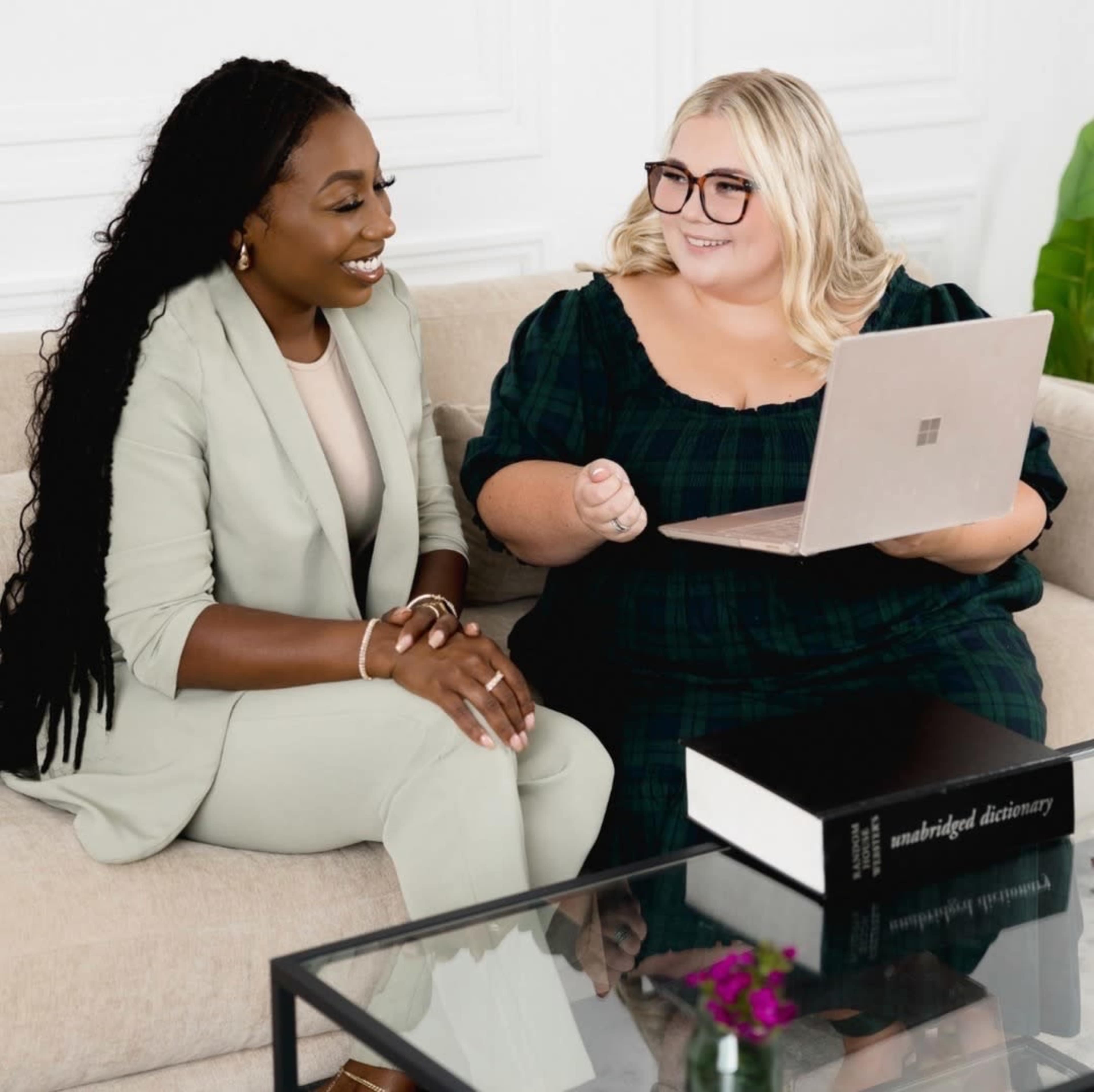 Two women sit on a sofa, one in a light-colored suit and the other in a dark plaid dress, while discussing a laptop on a coffee table.