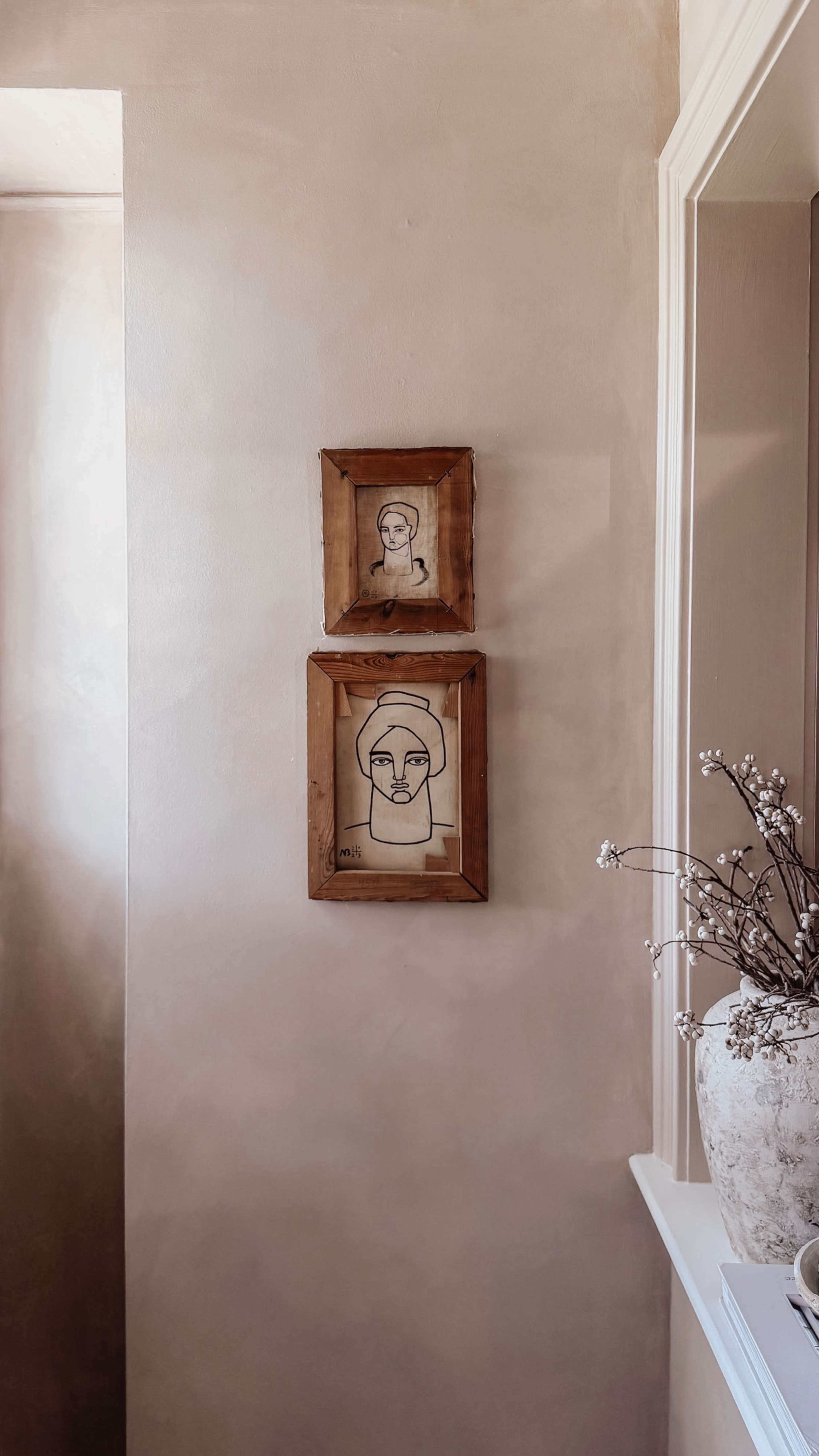 Two framed line art portraits hang on a light-colored wall, above a decorative vase with dried flowers.