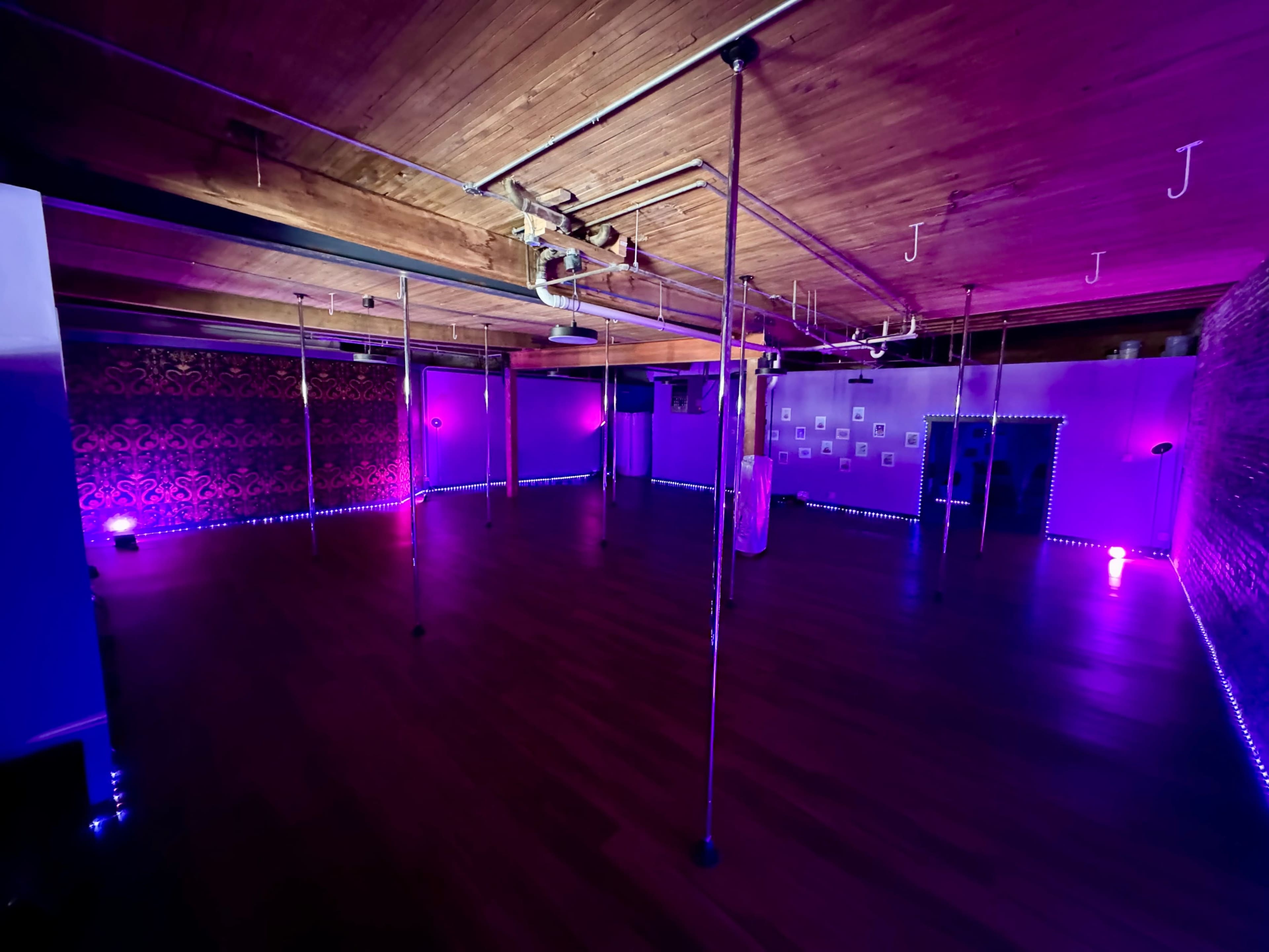 A spacious dance studio with polished wooden floors and multiple vertical poles, illuminated by purple and blue lighting.