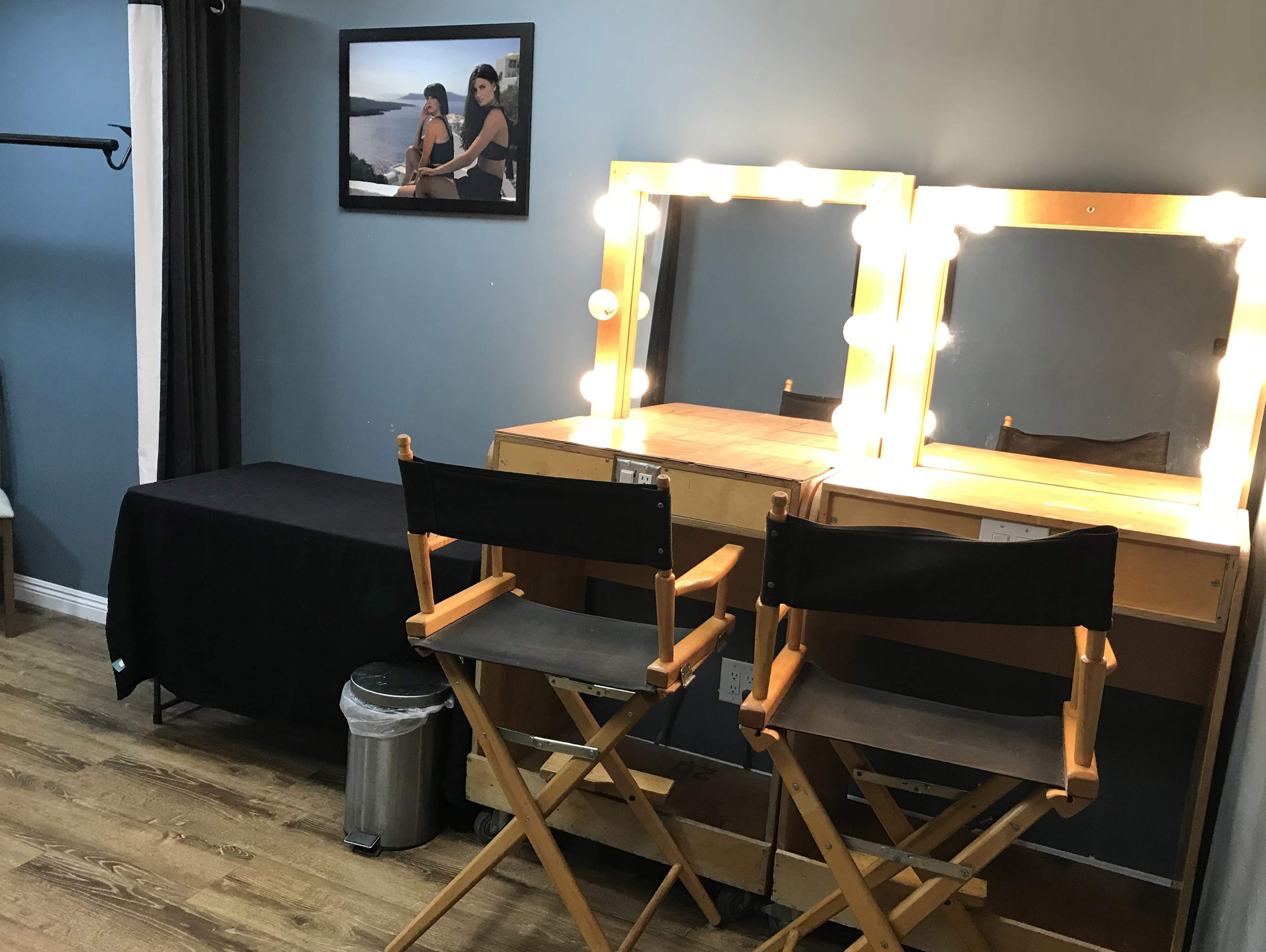 A makeup room featuring two illuminated vanity mirrors, a black table, and a photograph on the wall.