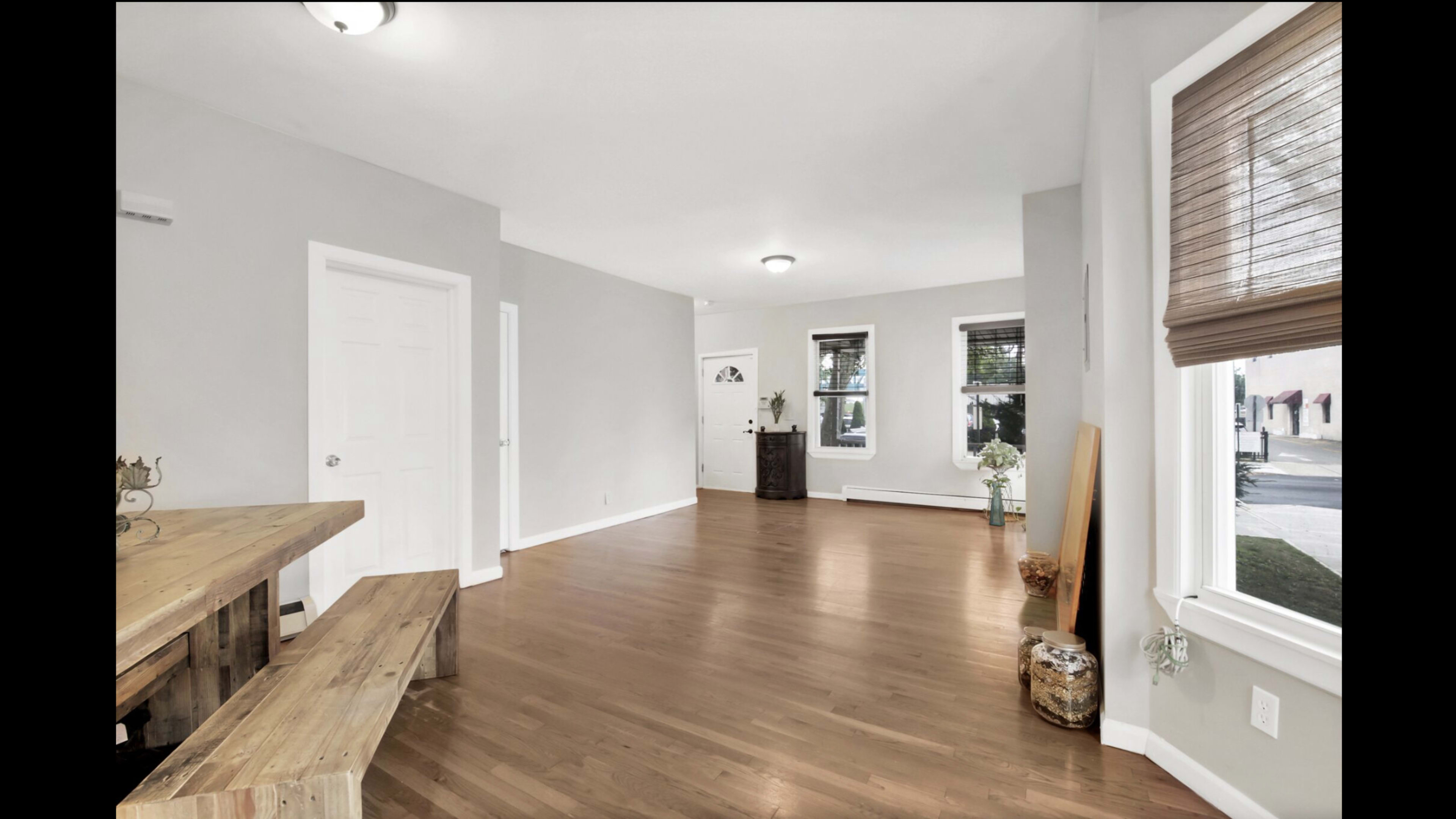 The image shows a spacious, light-colored interior room with wooden flooring, featuring a dining table and benches on one side and windows allowing natural light.