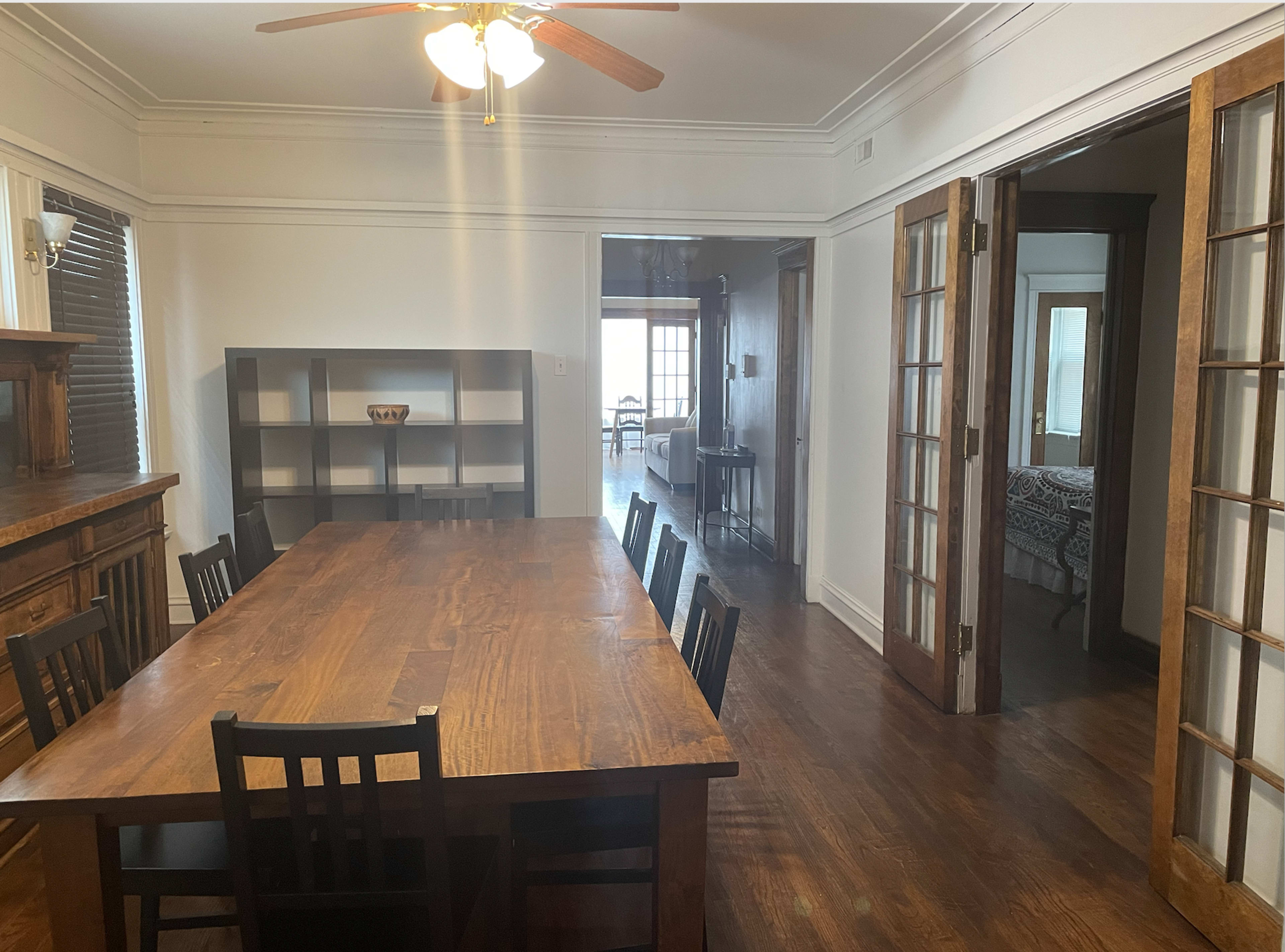 A large wooden dining table with black chairs is situated in a well-lit room, adjacent to an open doorway leading to another part of the house.