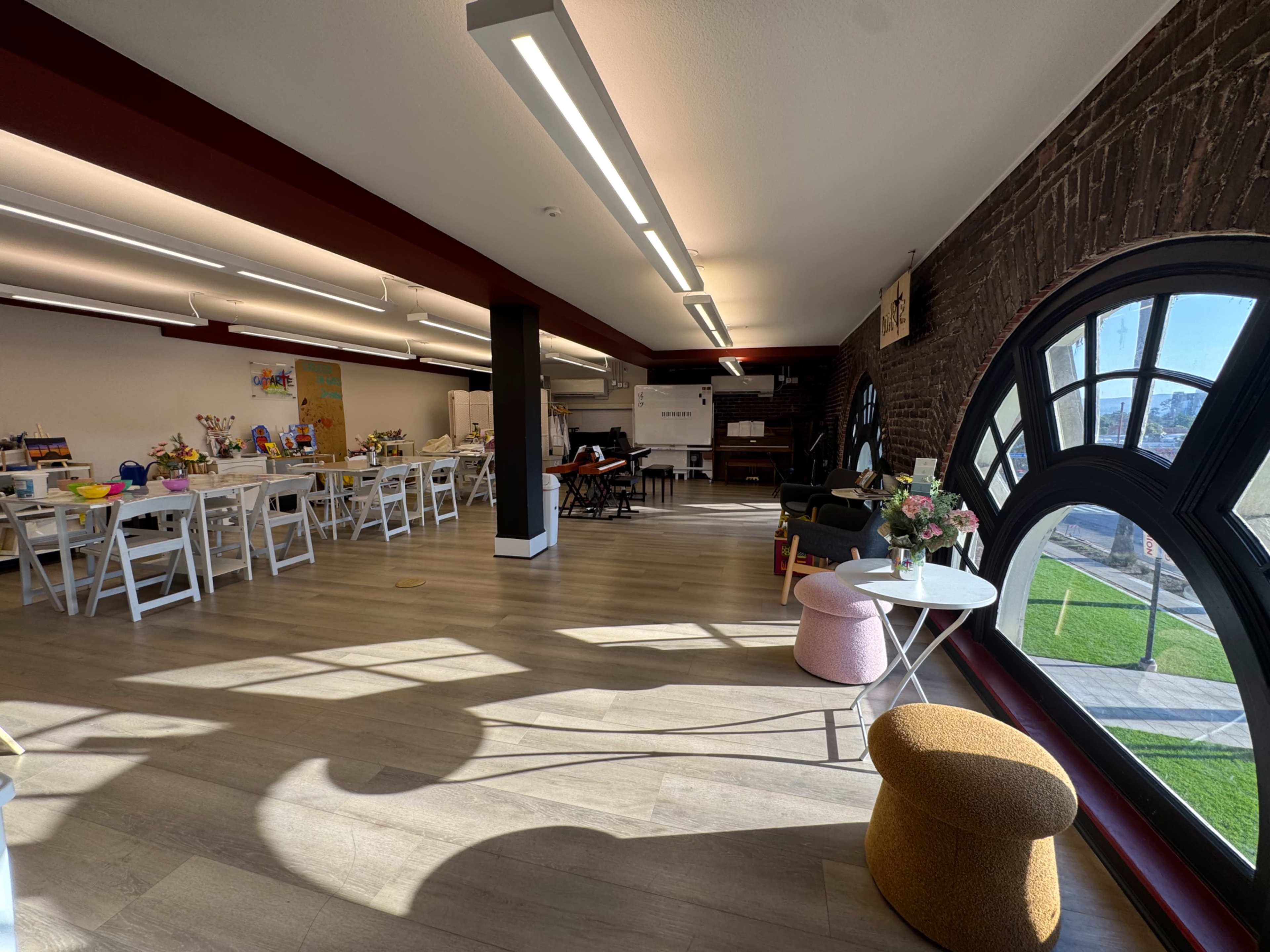 The image shows a spacious, well-lit community workspace featuring white tables, colorful seating options, and large windows that allow natural light to fill the room.
