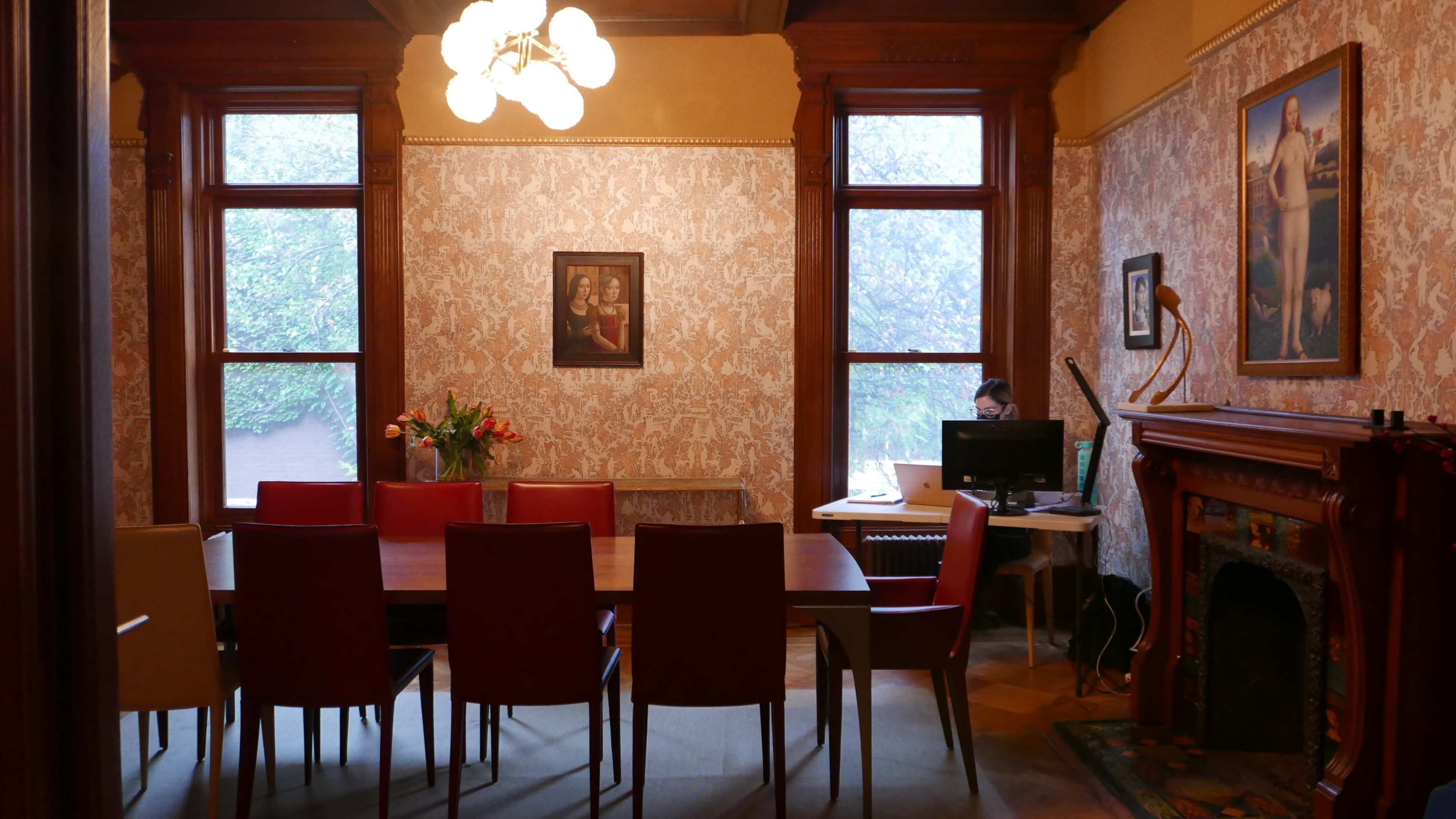 The image shows a room with a long dining table surrounded by chairs, a computer station in the corner, and vintage wallpaper decorated with portraits.