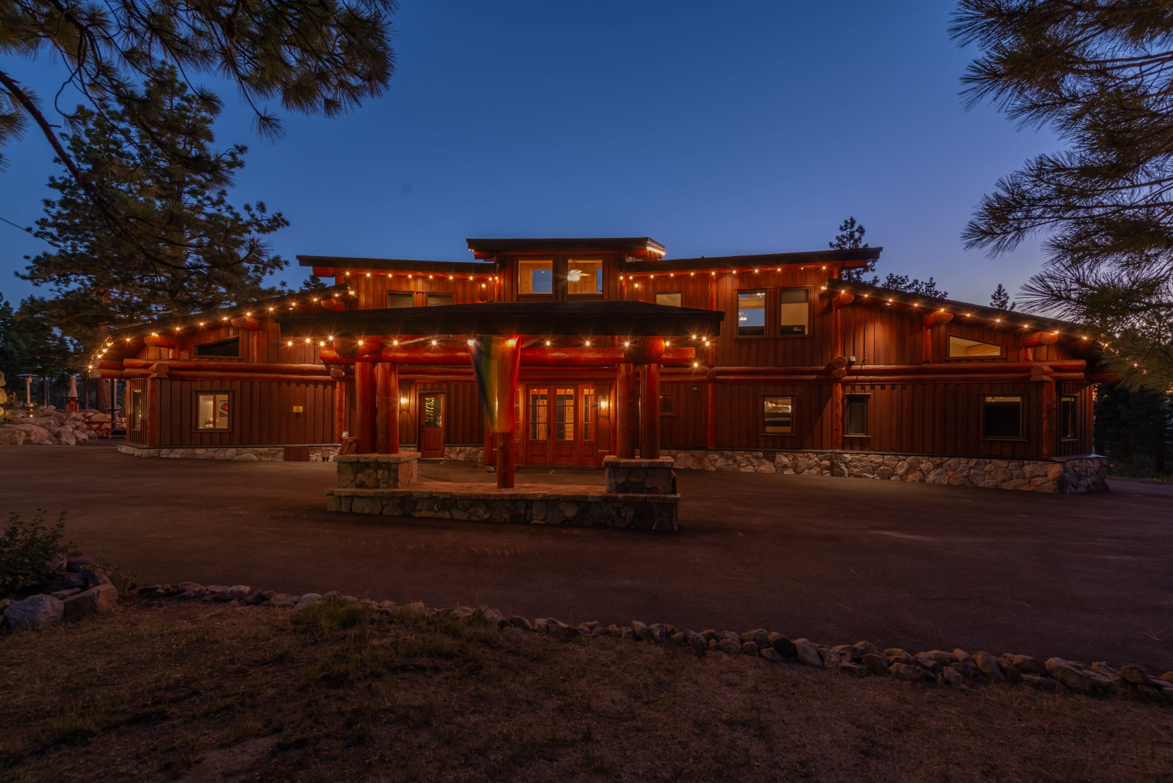 The image shows a large, illuminated wooden lodge with a spacious entrance surrounded by trees at twilight.