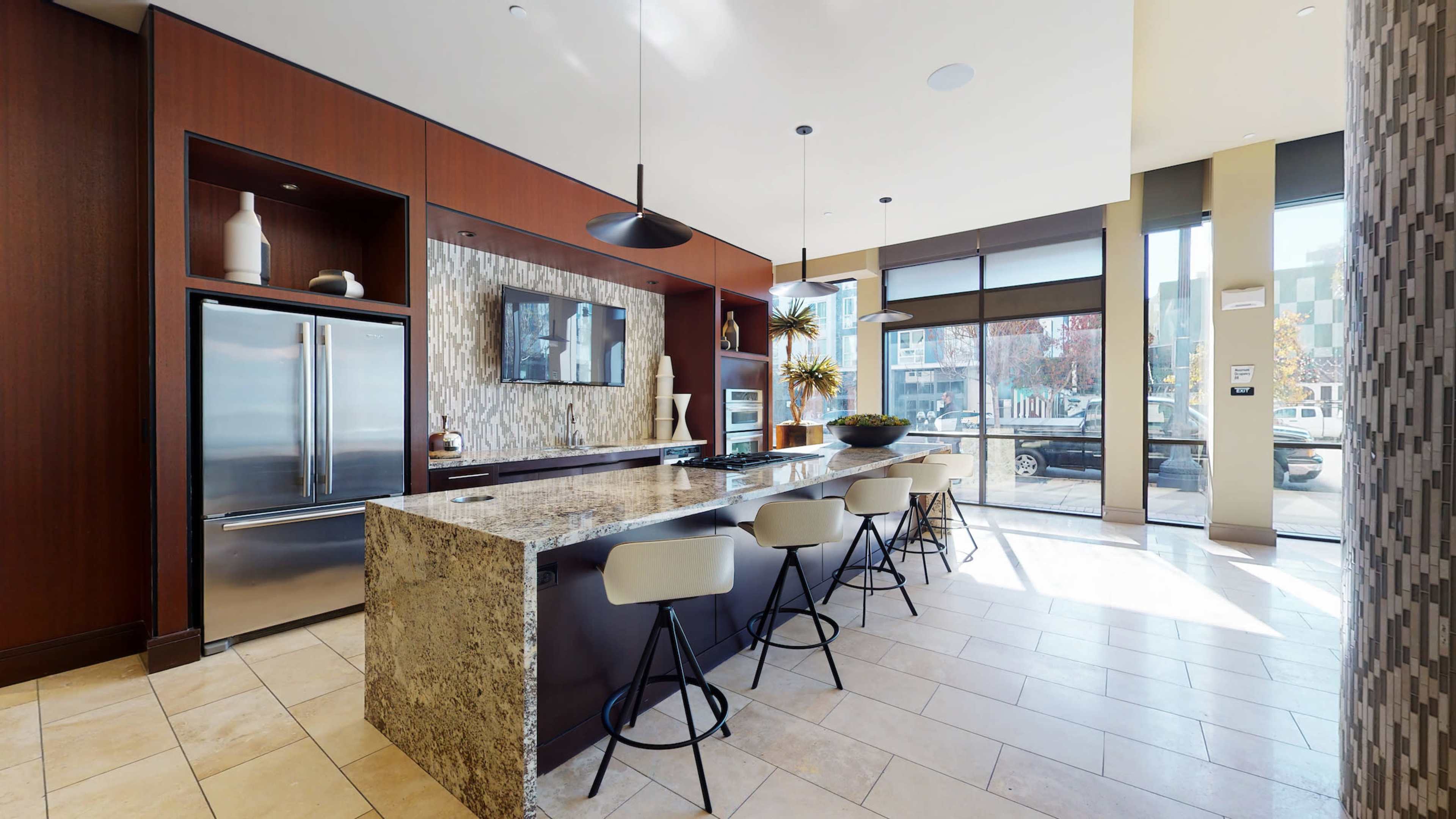 The image shows a modern kitchen with a large stone island, bar stools, stainless steel appliances, and windows that let in natural light.