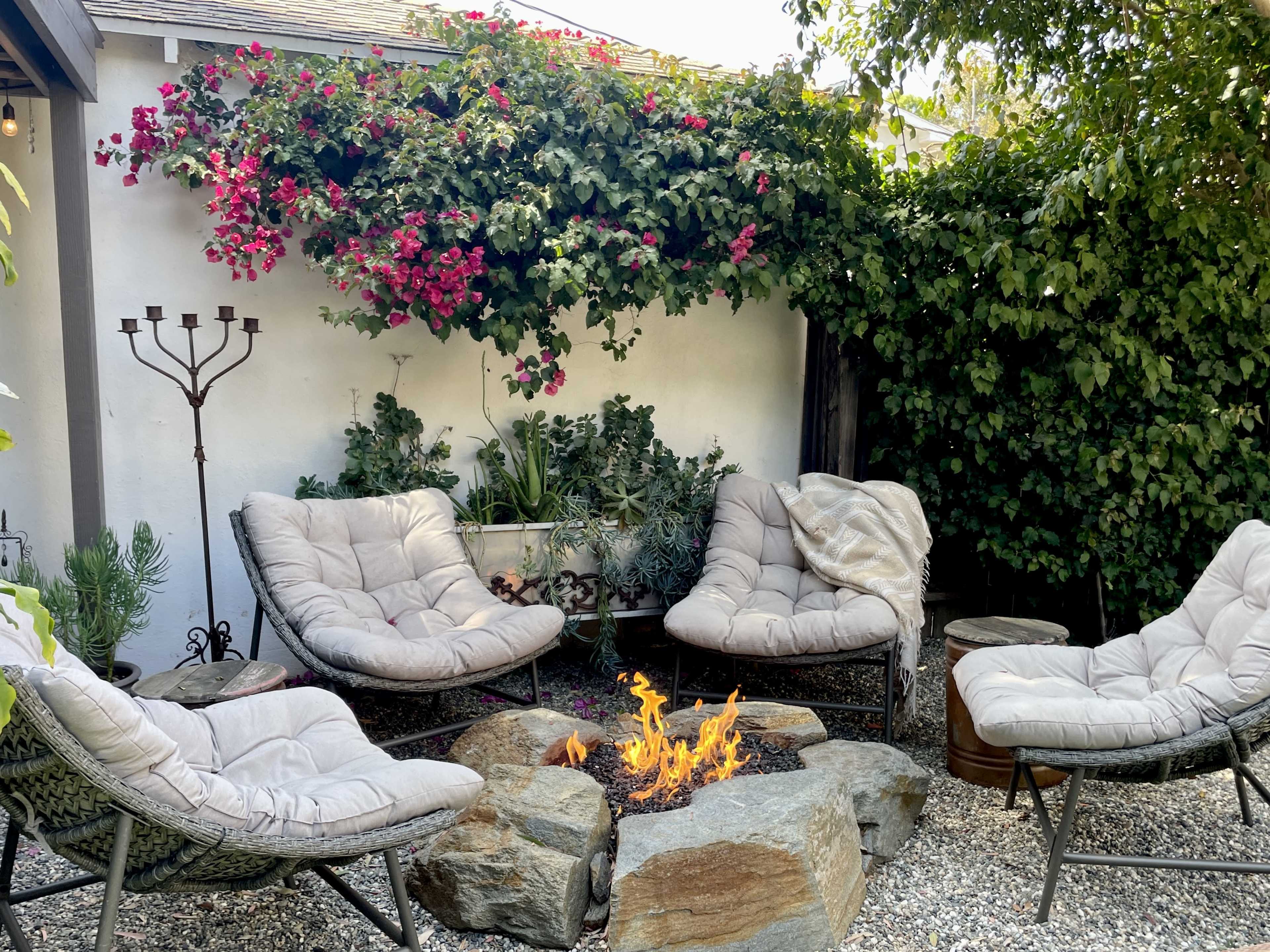 The image shows a cozy outdoor seating area surrounded by greenery, featuring four cushioned chairs arranged around a small fire pit made of rocks.