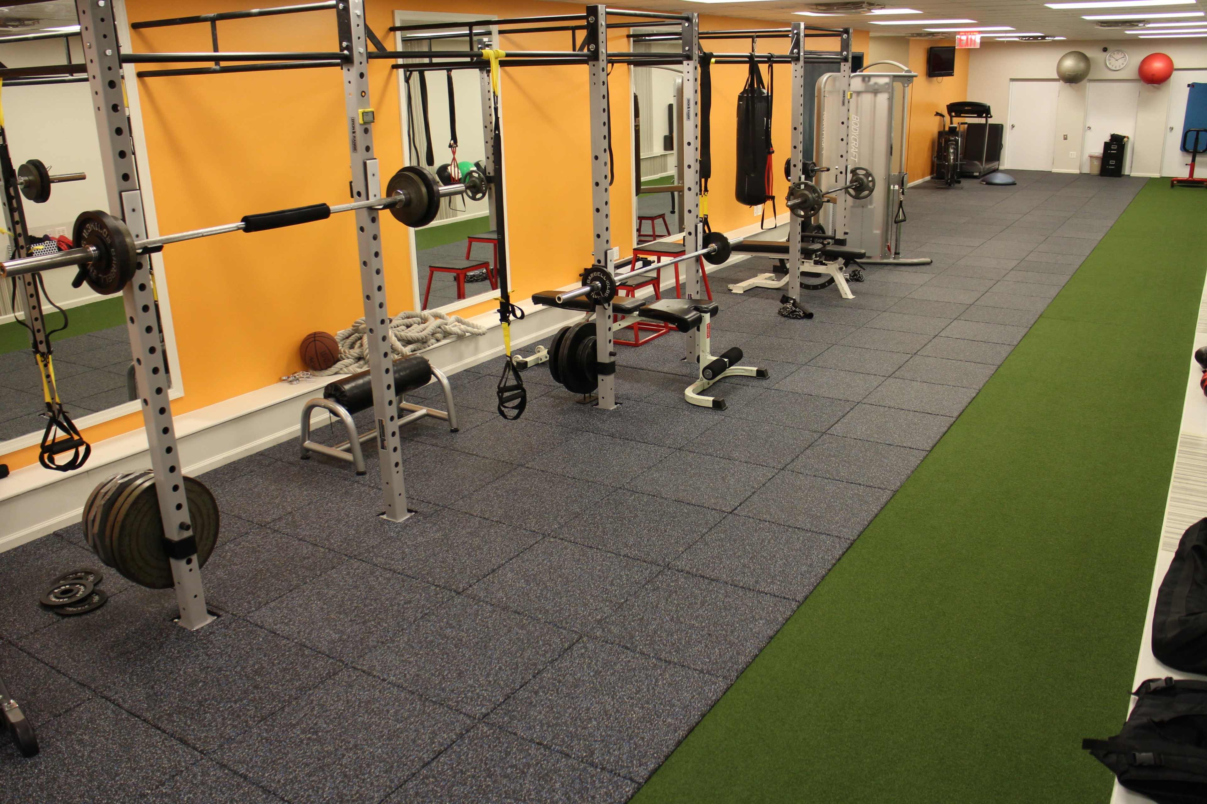 The image shows a well-equipped gym area with a variety of exercise equipment, including weightlifting bars, benches, and mats on a green turf floor.