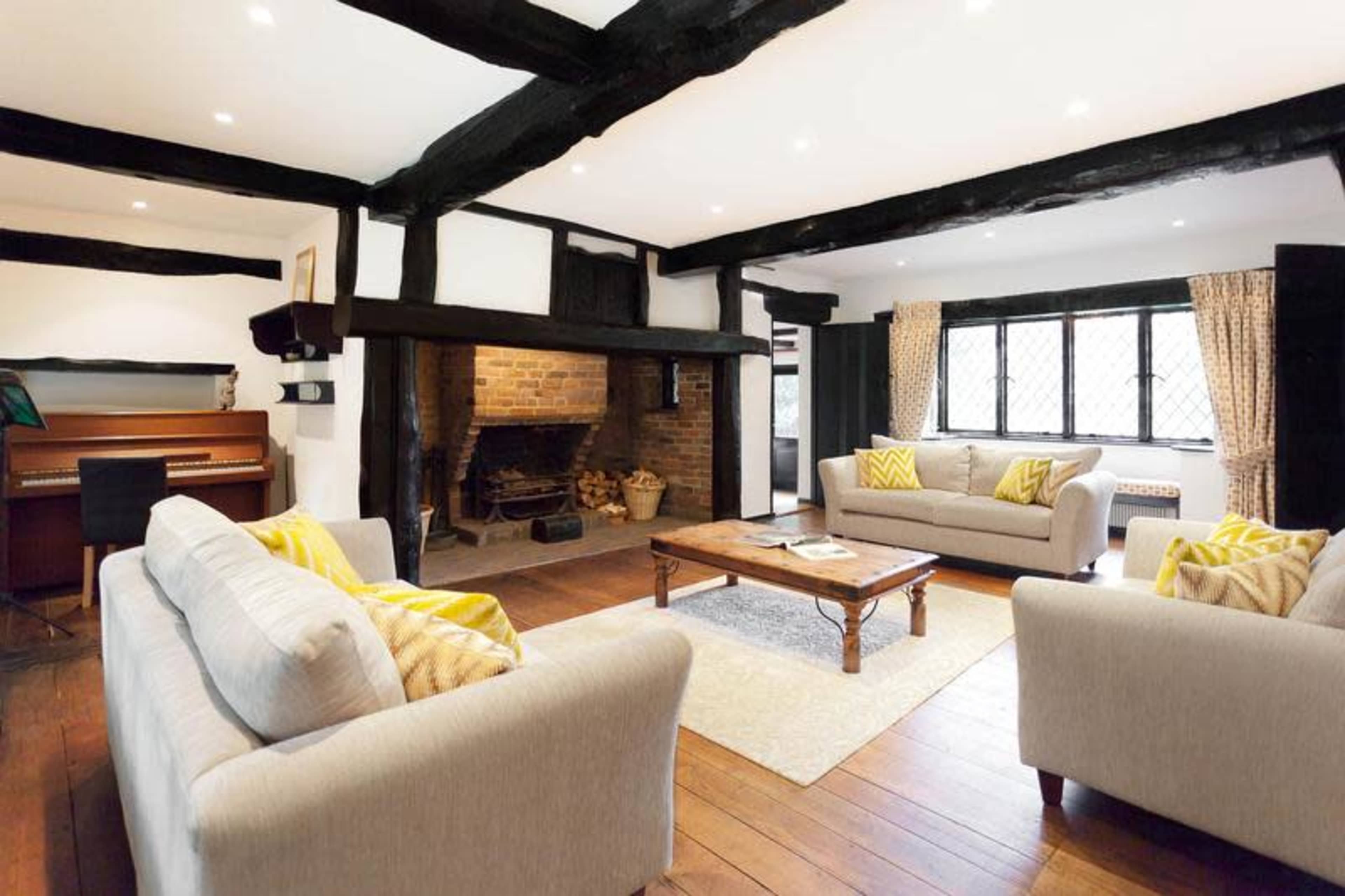 A spacious living room features two light-colored sofas, a wooden coffee table, a brick fireplace, and a piano in a cozy setting with exposed beams and large windows.