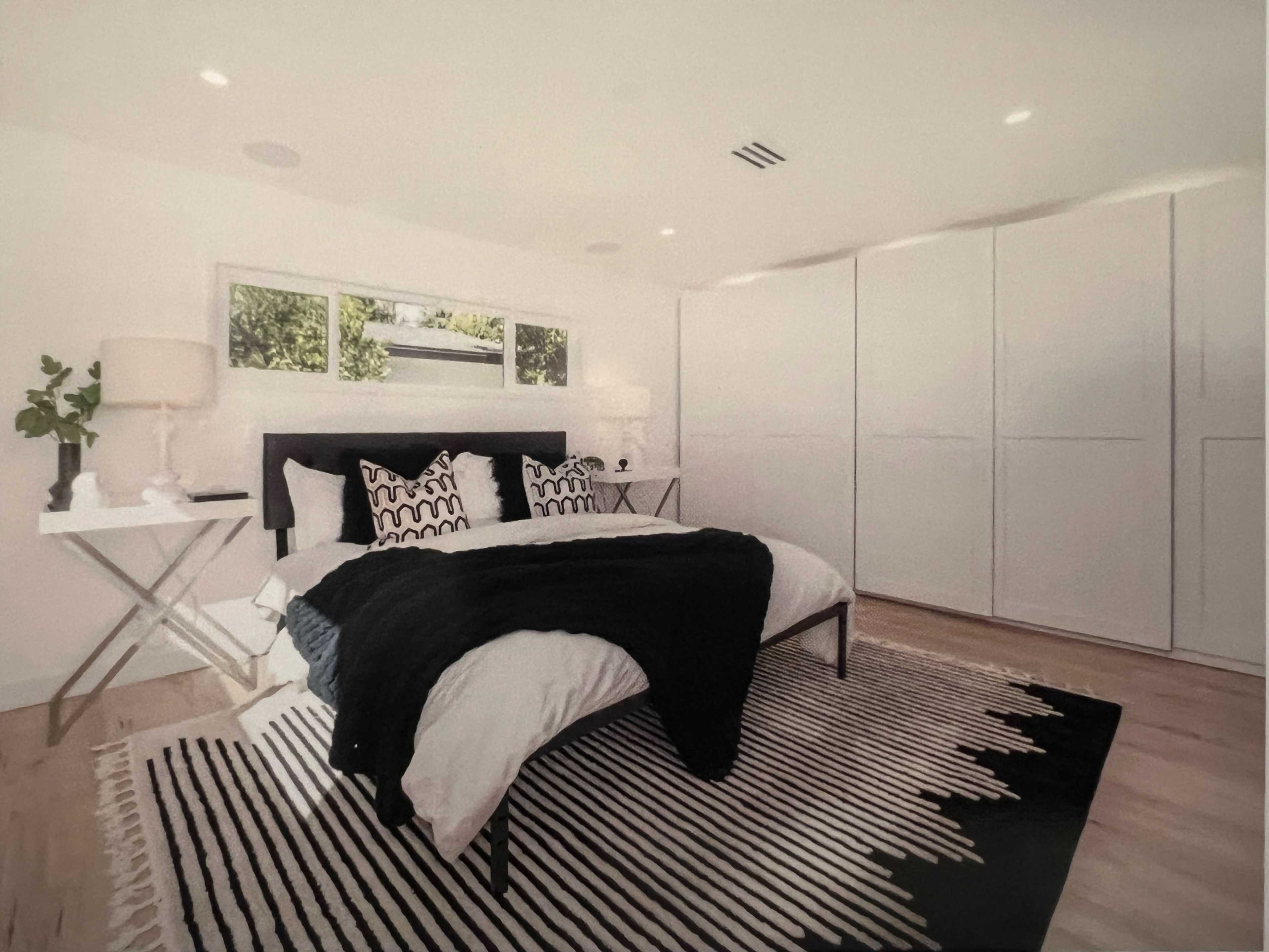 The image depicts a modern bedroom featuring a bed with dark bedding, a striped rug, a side table with a lamp, and large white wardrobes.