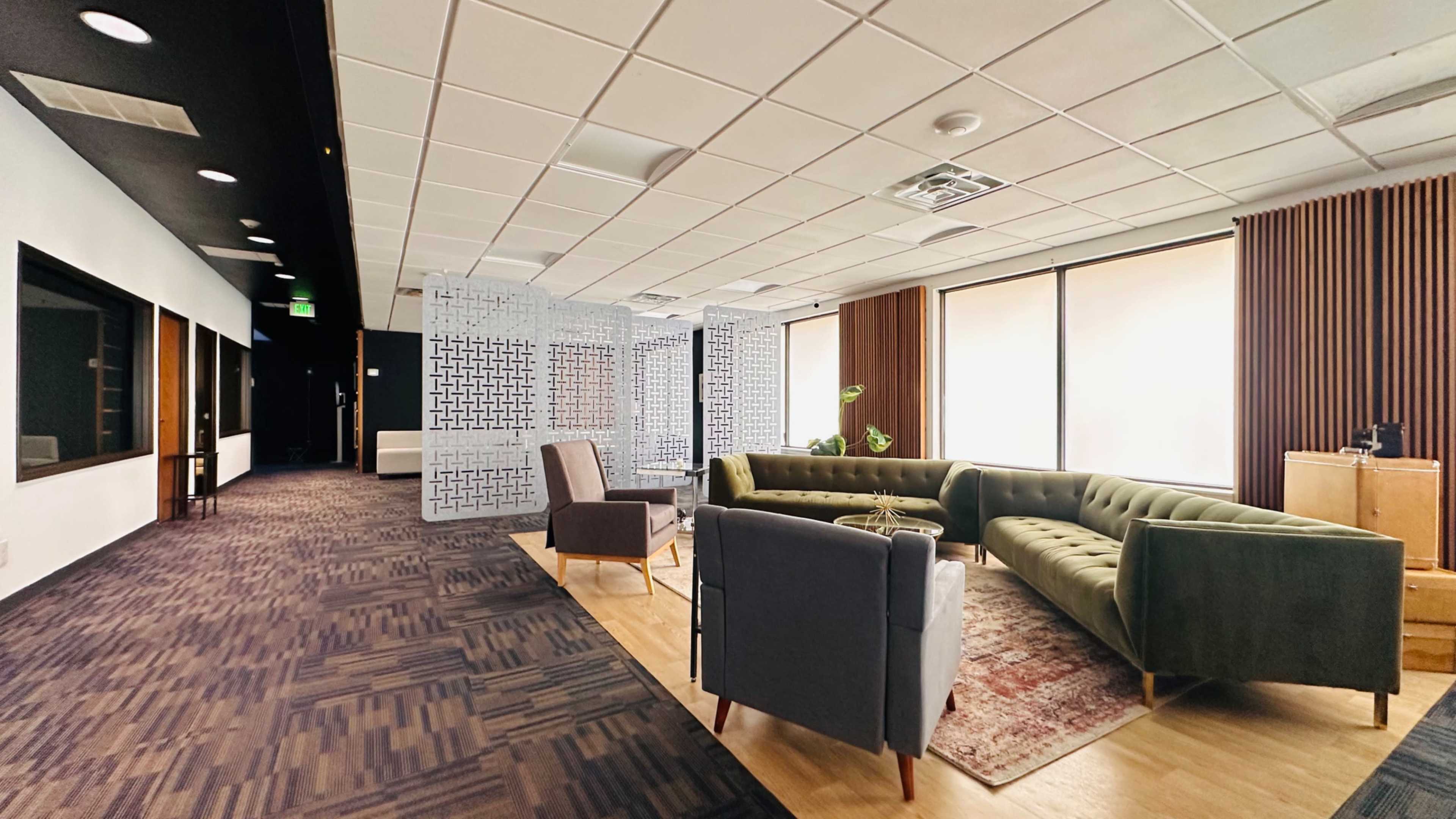 The image shows a modern office interior featuring a green sectional sofa, a gray armchair, and large windows with soft natural light.