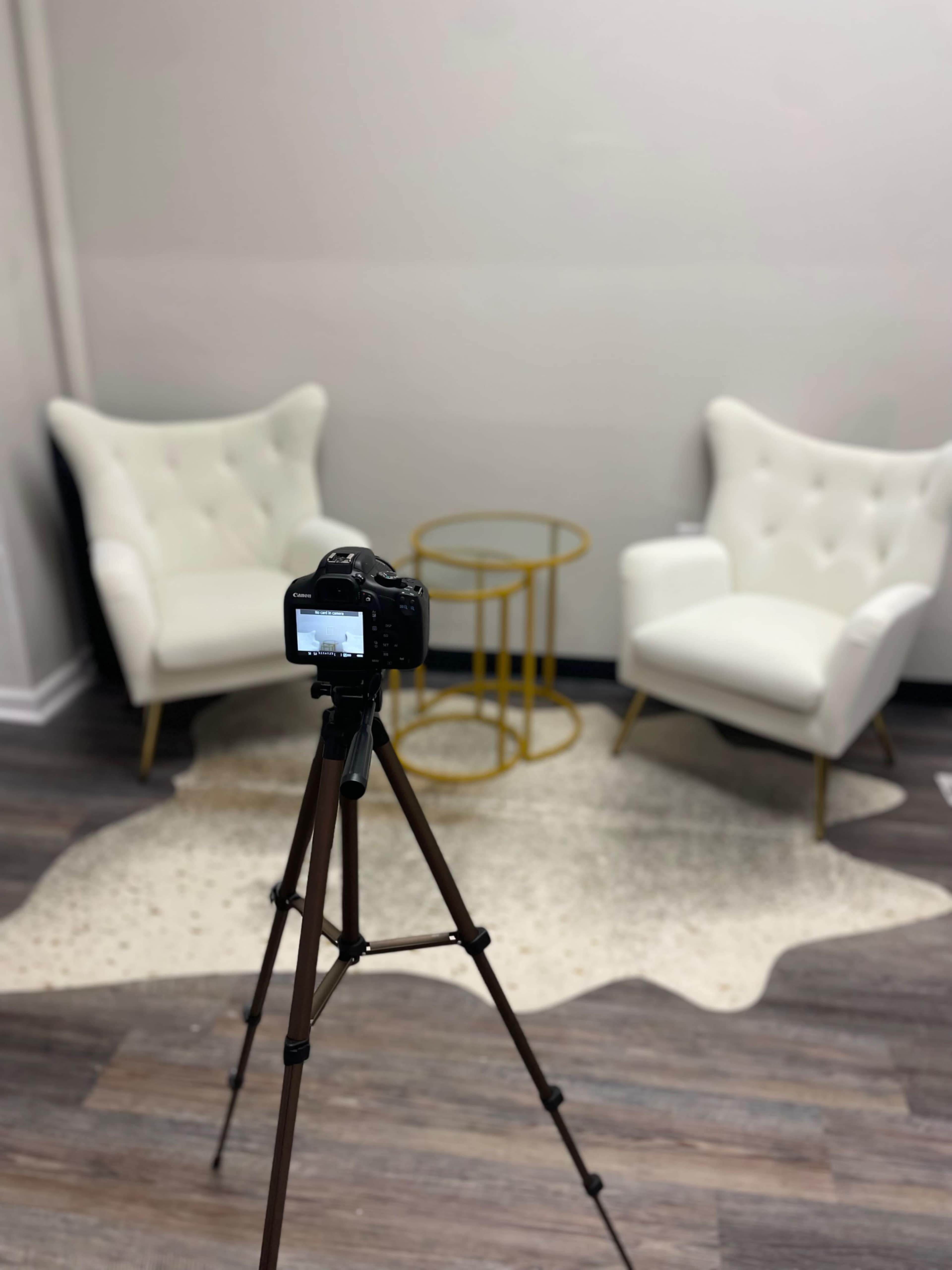 A camera on a tripod faces two white armchairs and a glass side table in a simple interior setting.