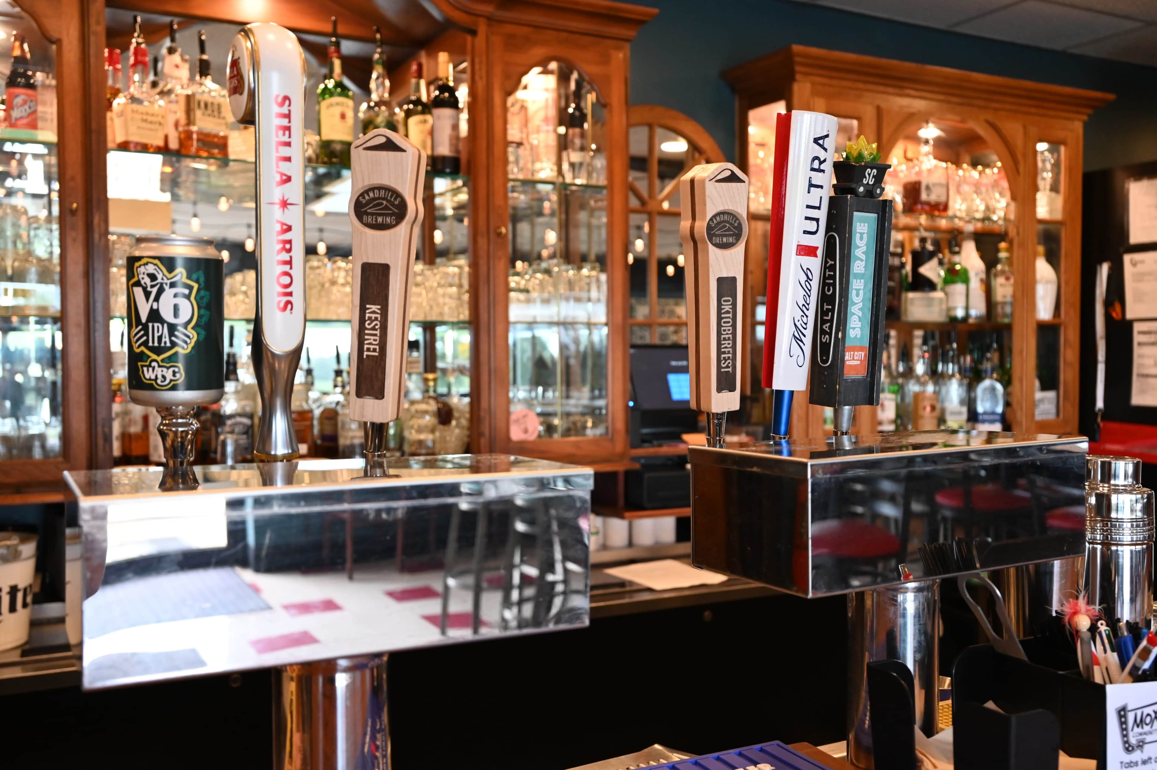 The image shows a bar area with several beer taps, including brands like Stella Artois and Budweiser, in front of a mirrored backdrop displaying various liquor bottles.
