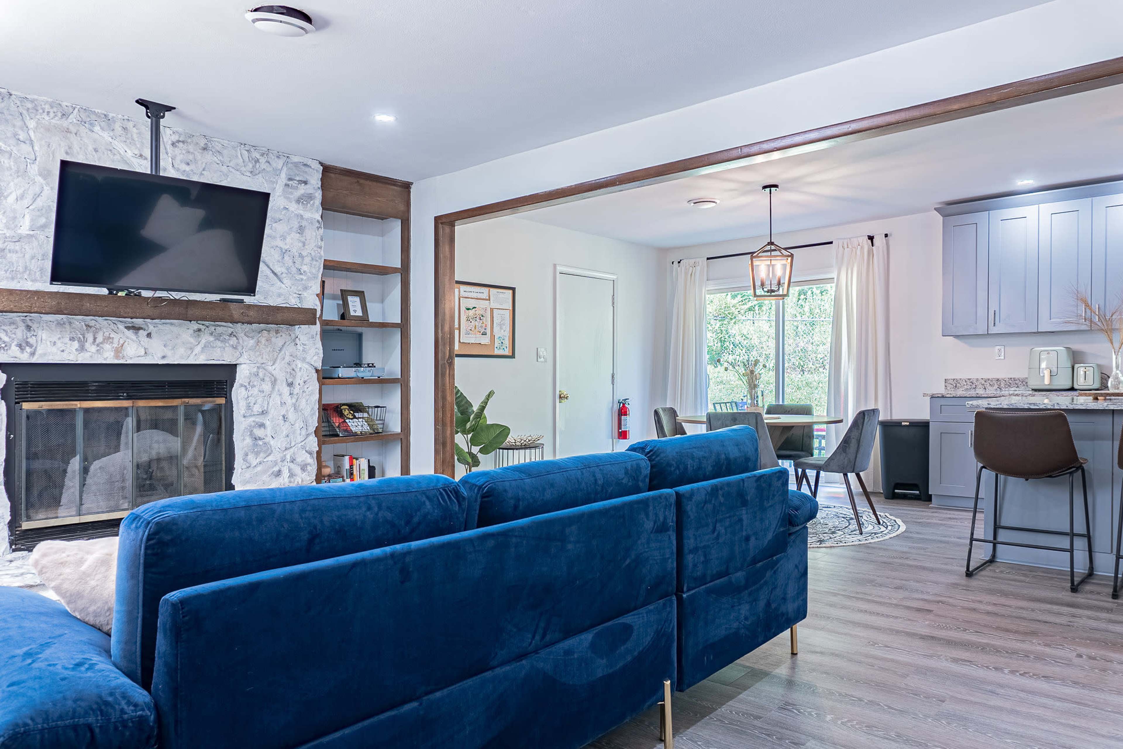 A modern living room with a blue sectional sofa, a stone fireplace, and an open layout leading to a dining area with hardwood floors and a kitchen in the background.