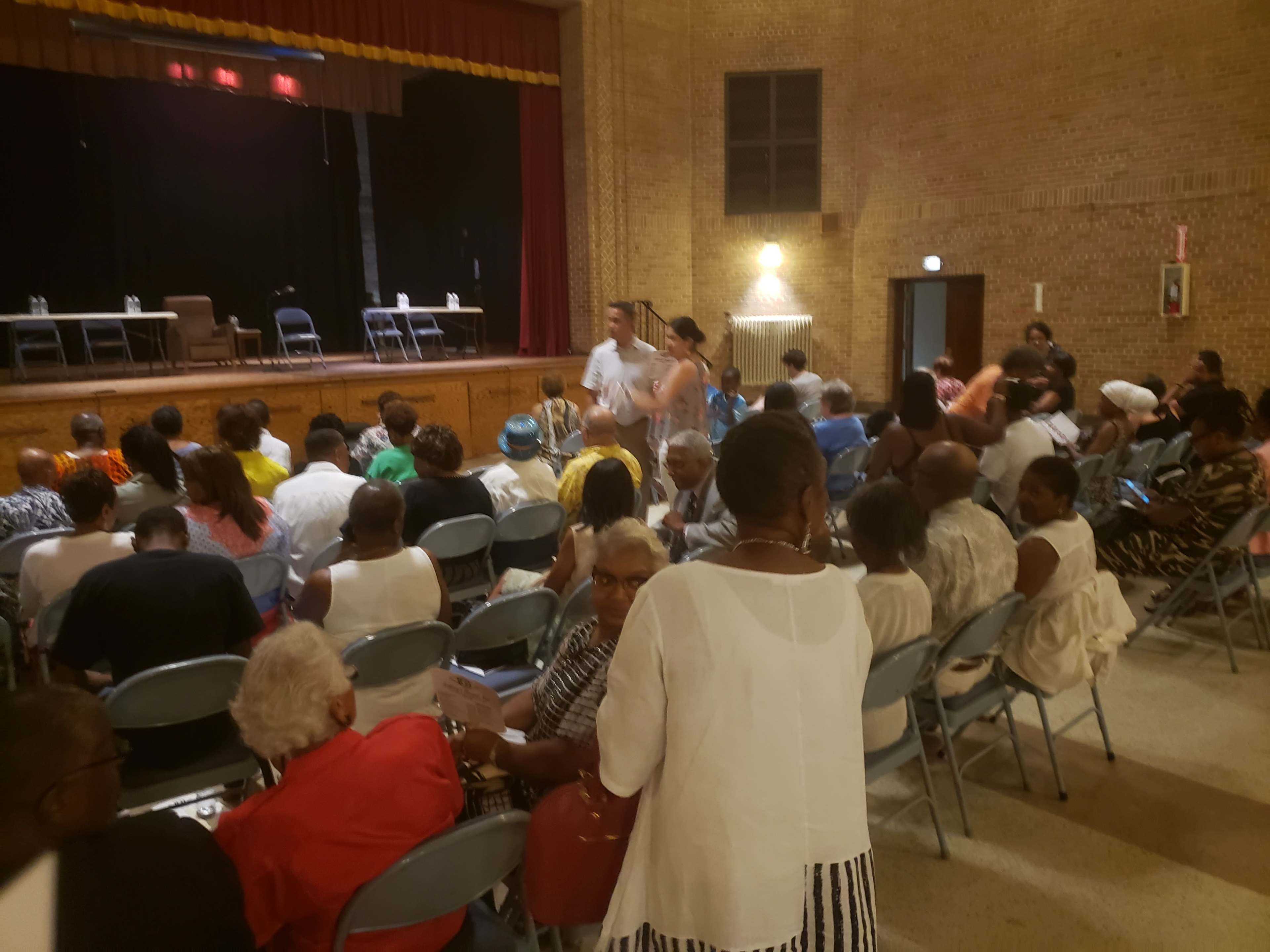 A diverse audience is seated in a community auditorium, facing a stage with tables and chairs positioned for an event.