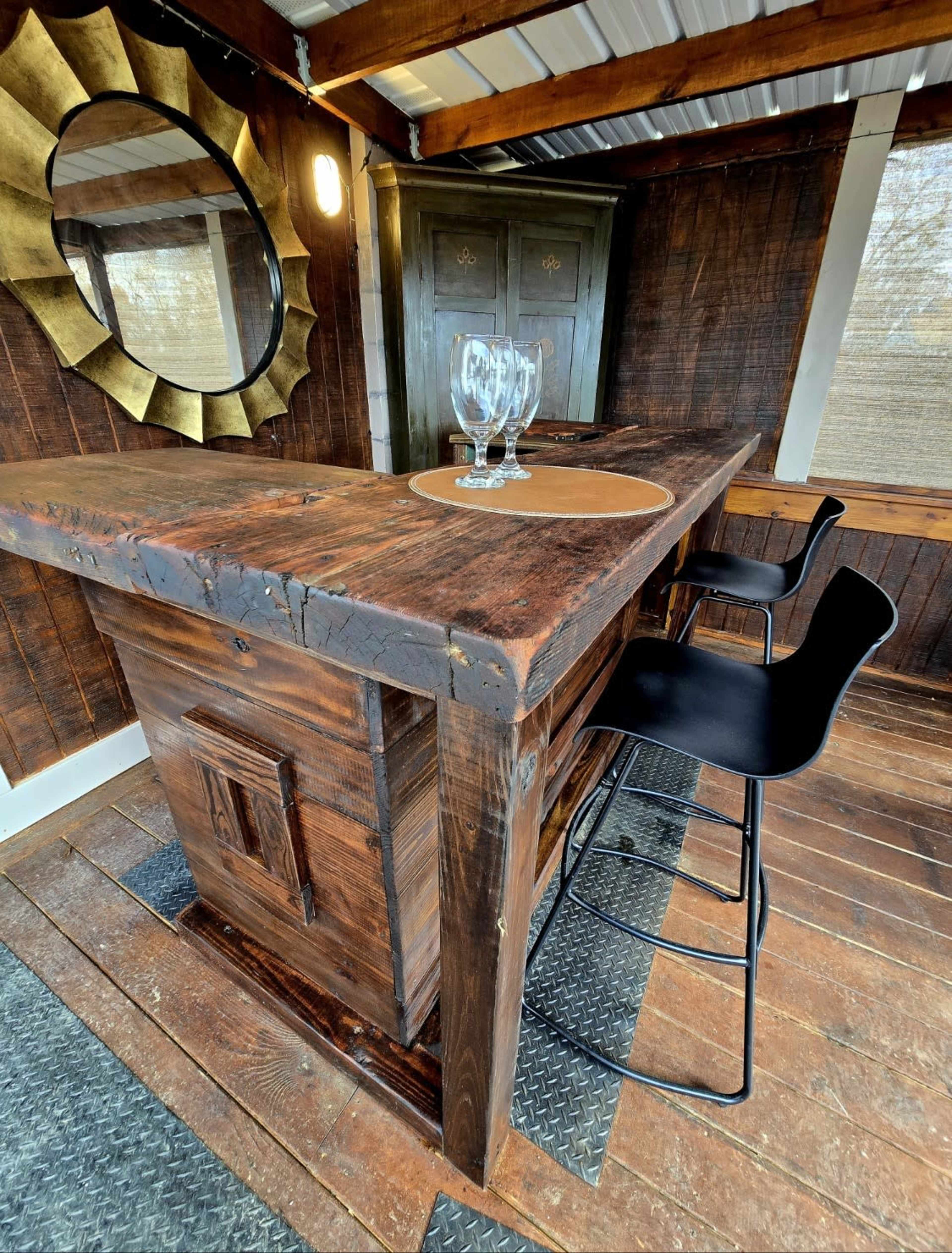 The image shows a rustic wooden bar with two black stools, a round placemat, and two clear glasses on top, set within a cozy indoor space featuring dark wooden walls.