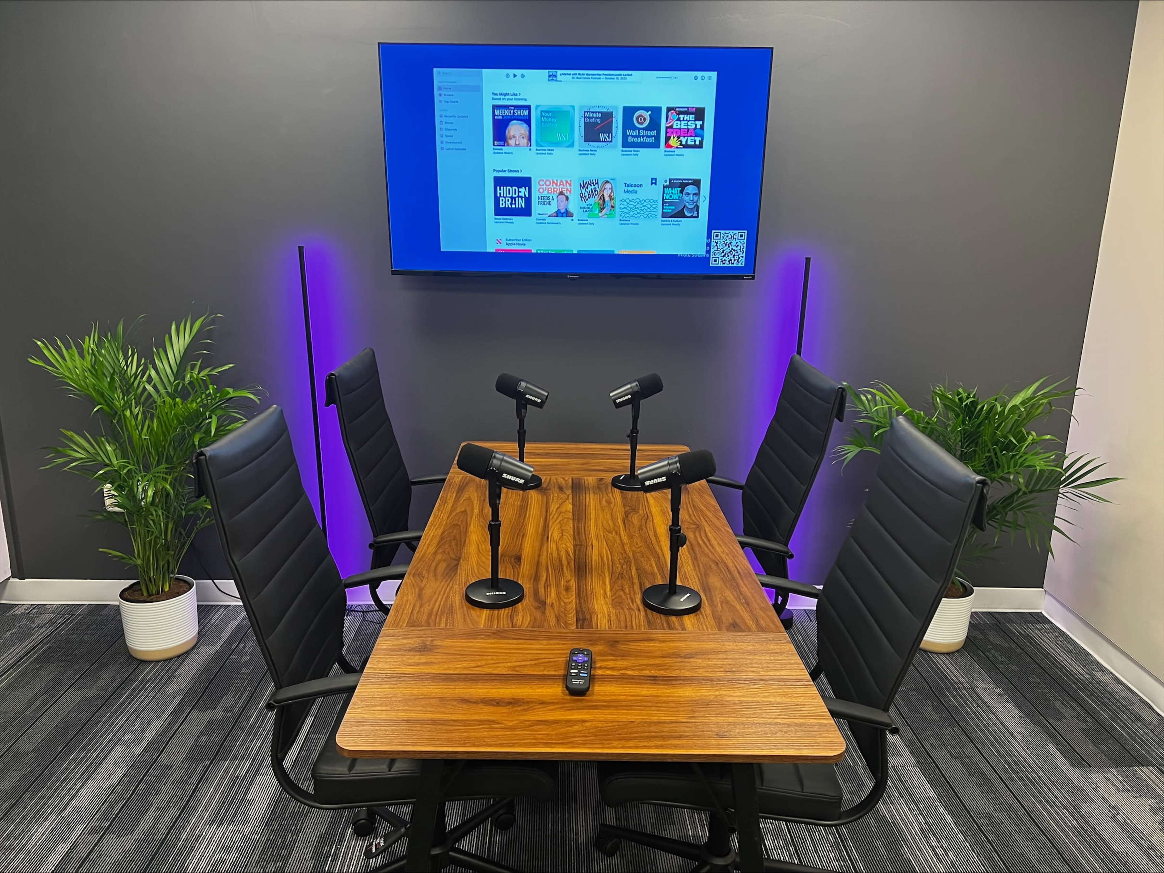 A modern conference room features a wooden table surrounded by black chairs, with four microphones positioned for use and a large screen displaying digital content in the background.