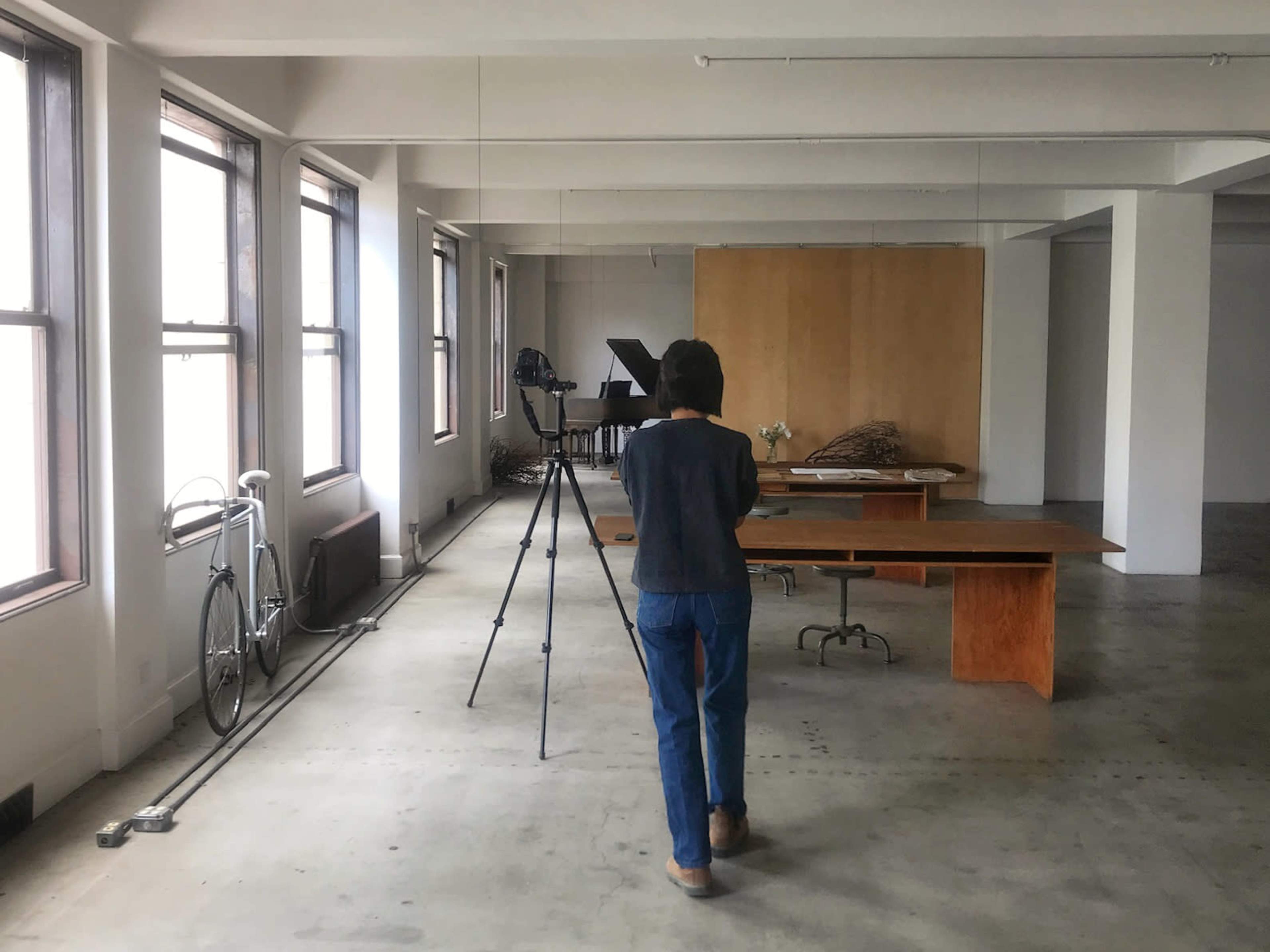 A person stands in a spacious, minimalist room with large windows, a wooden table, and a bicycle against the wall.