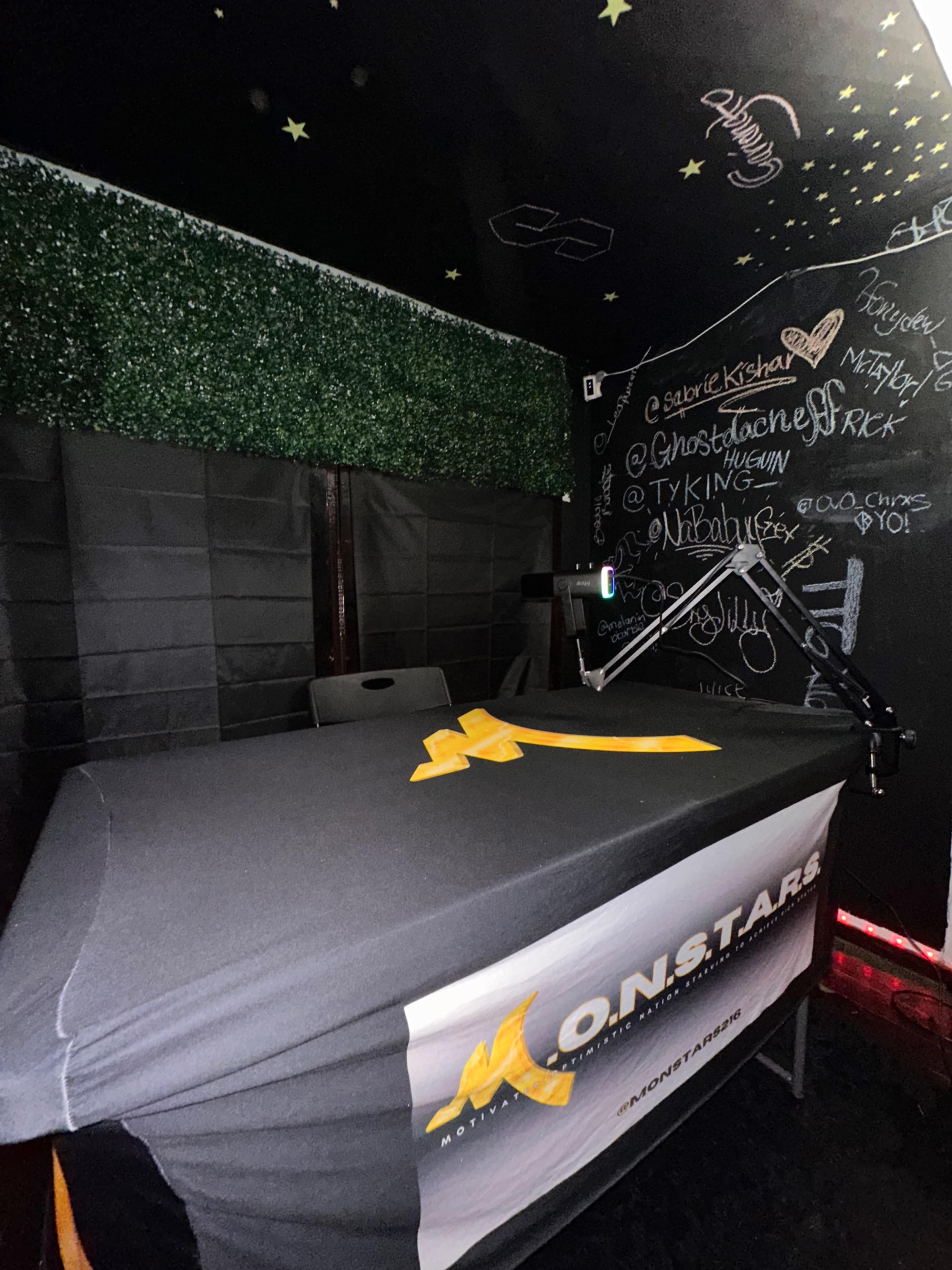 The image shows a partially enclosed space with a table covered by a black and orange tablecloth, featuring the text "M.O.N.STARS," and a microphone mounted on an arm.