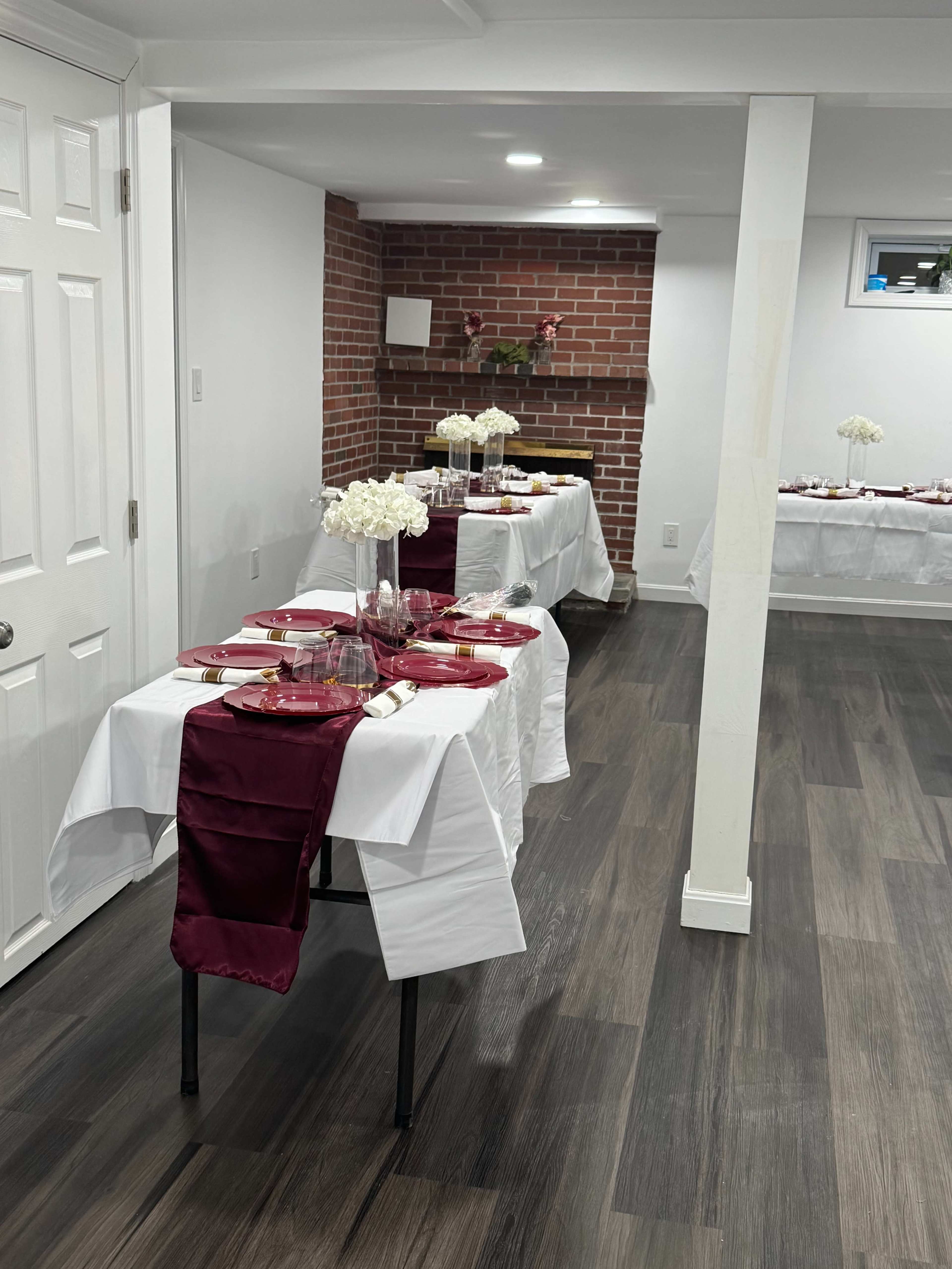 The image shows a decorative dining setup in a basement with tables covered in white tablecloths, each featuring burgundy runners, plates, and floral centerpieces.