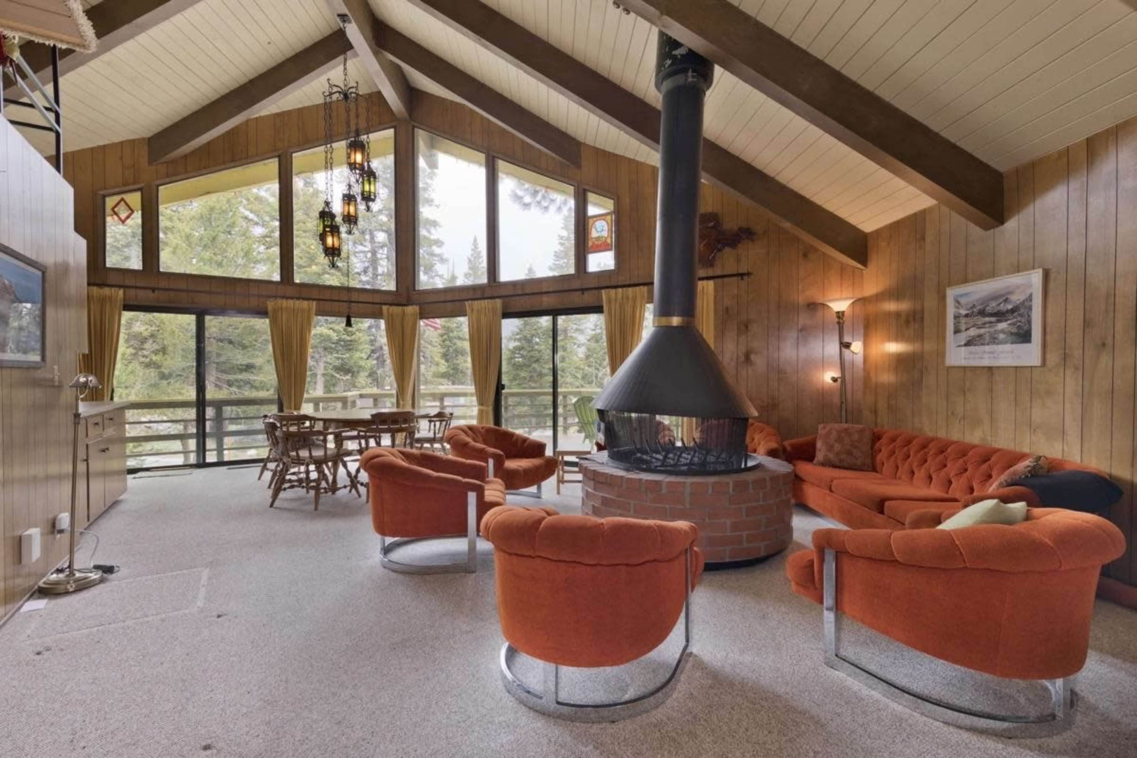 A cozy living room features a central circular fireplace, surrounded by orange furniture and large windows that provide views of the trees outside.