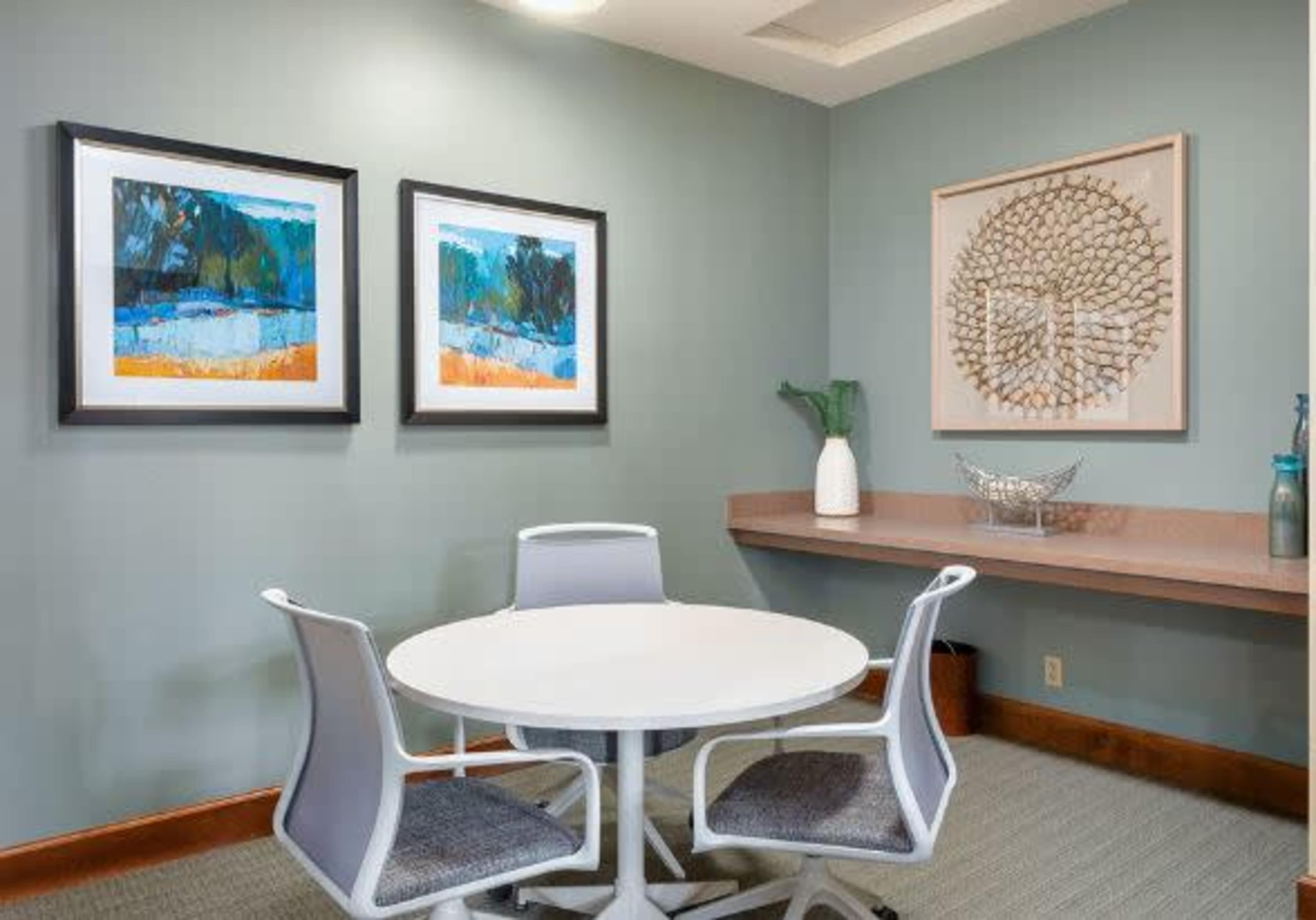 A small meeting room featuring a round white table surrounded by four chairs, with two framed artworks and a decorative wall panel.