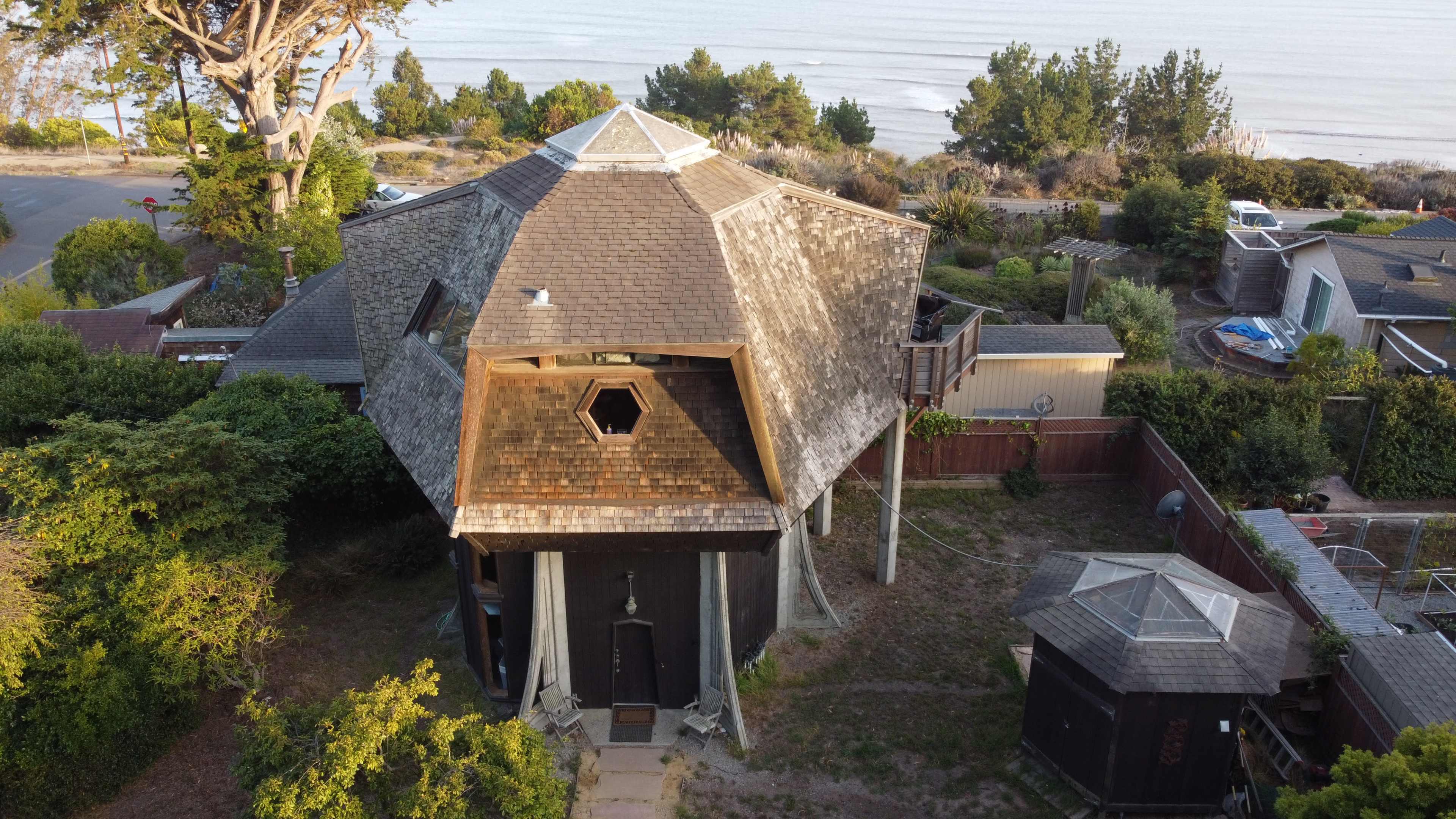An octagonal house with a central skylight stands surrounded by greenery and overlooks a beach in the background.