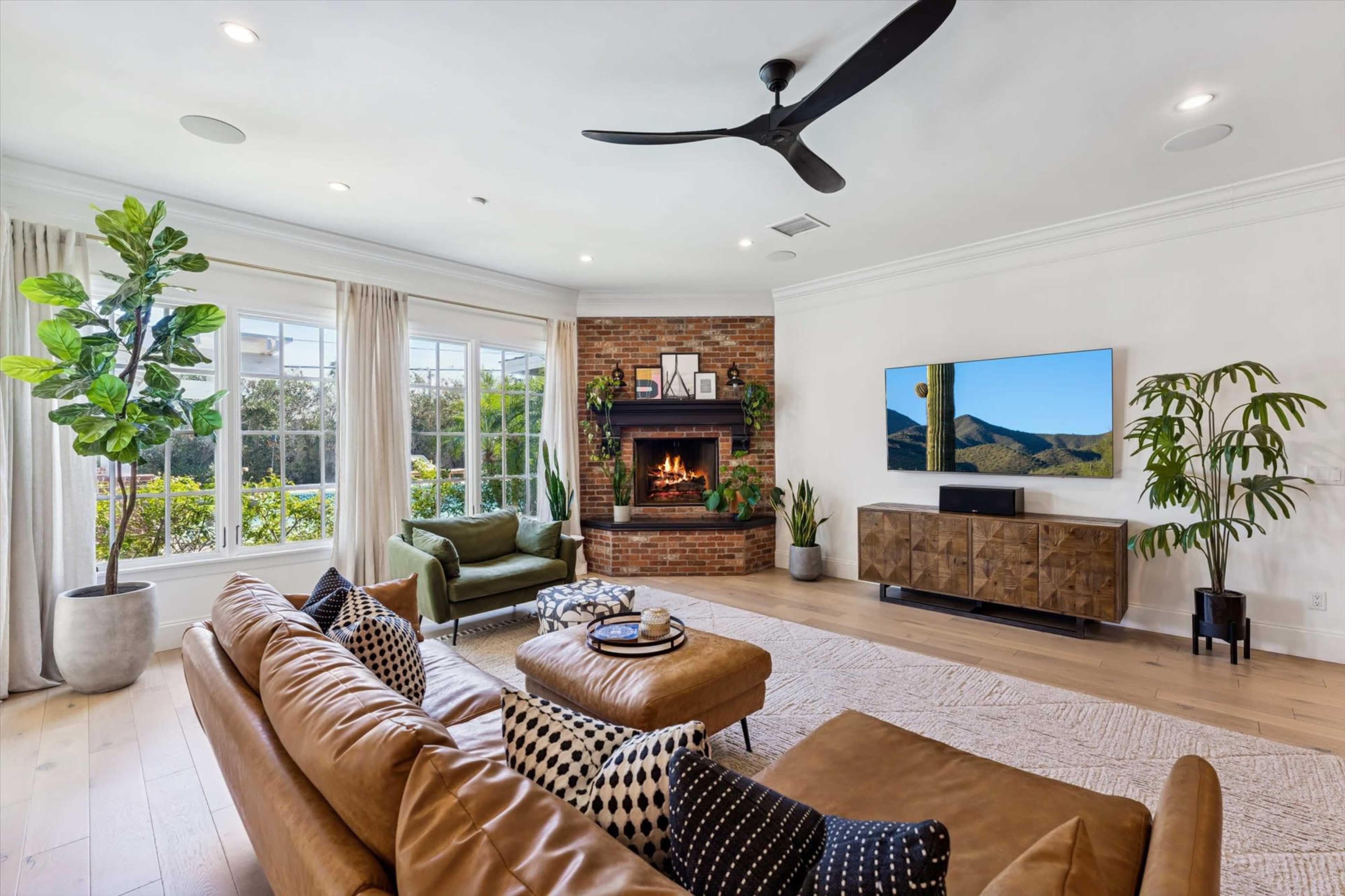 The living room features a leather sectional sofa, a modern fireplace with a TV above, large windows, and decorative plants.