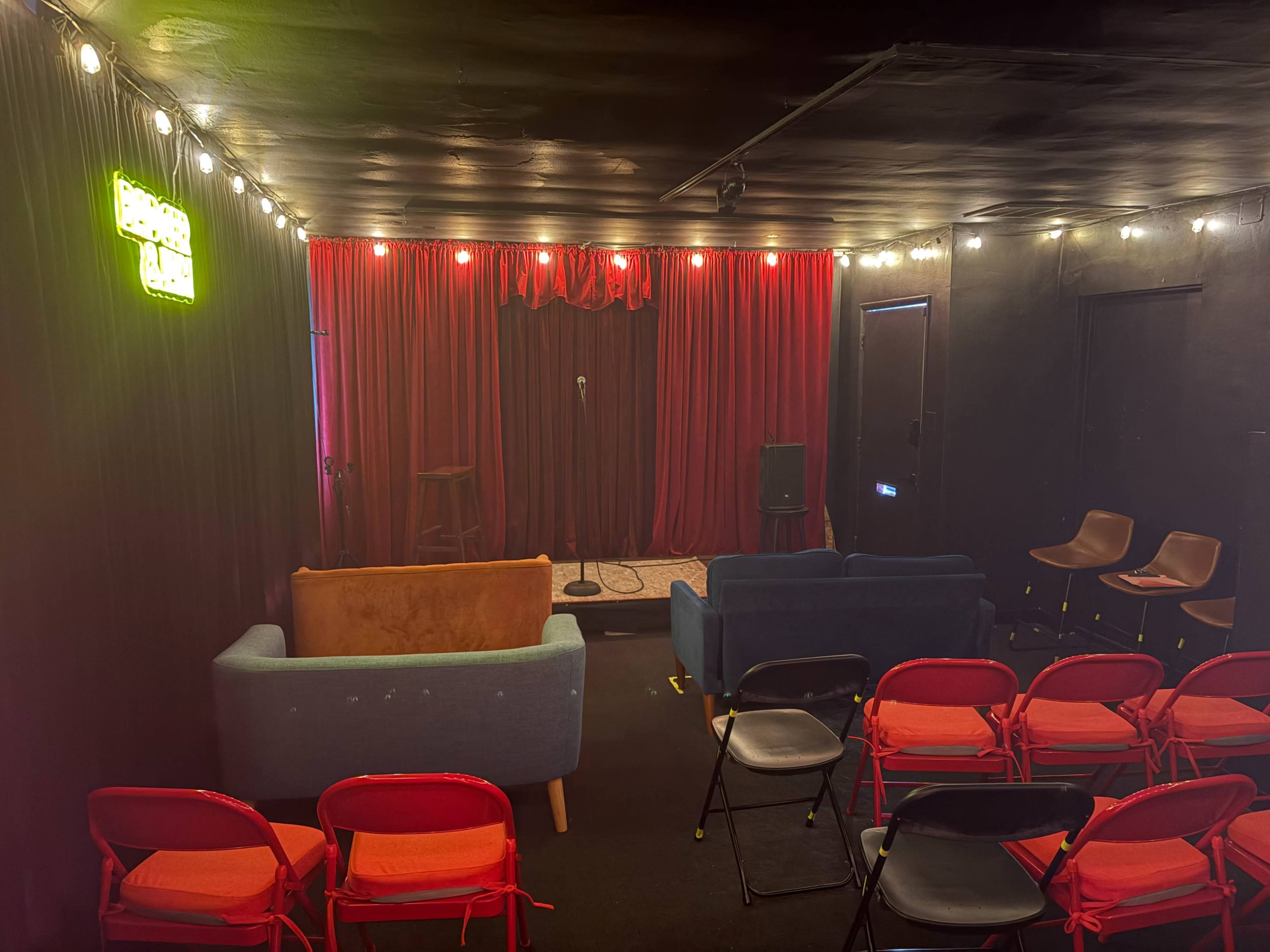 A small performance venue features a stage with a microphone, surrounded by red curtains, and various seating arrangements including red chairs and sofas.
