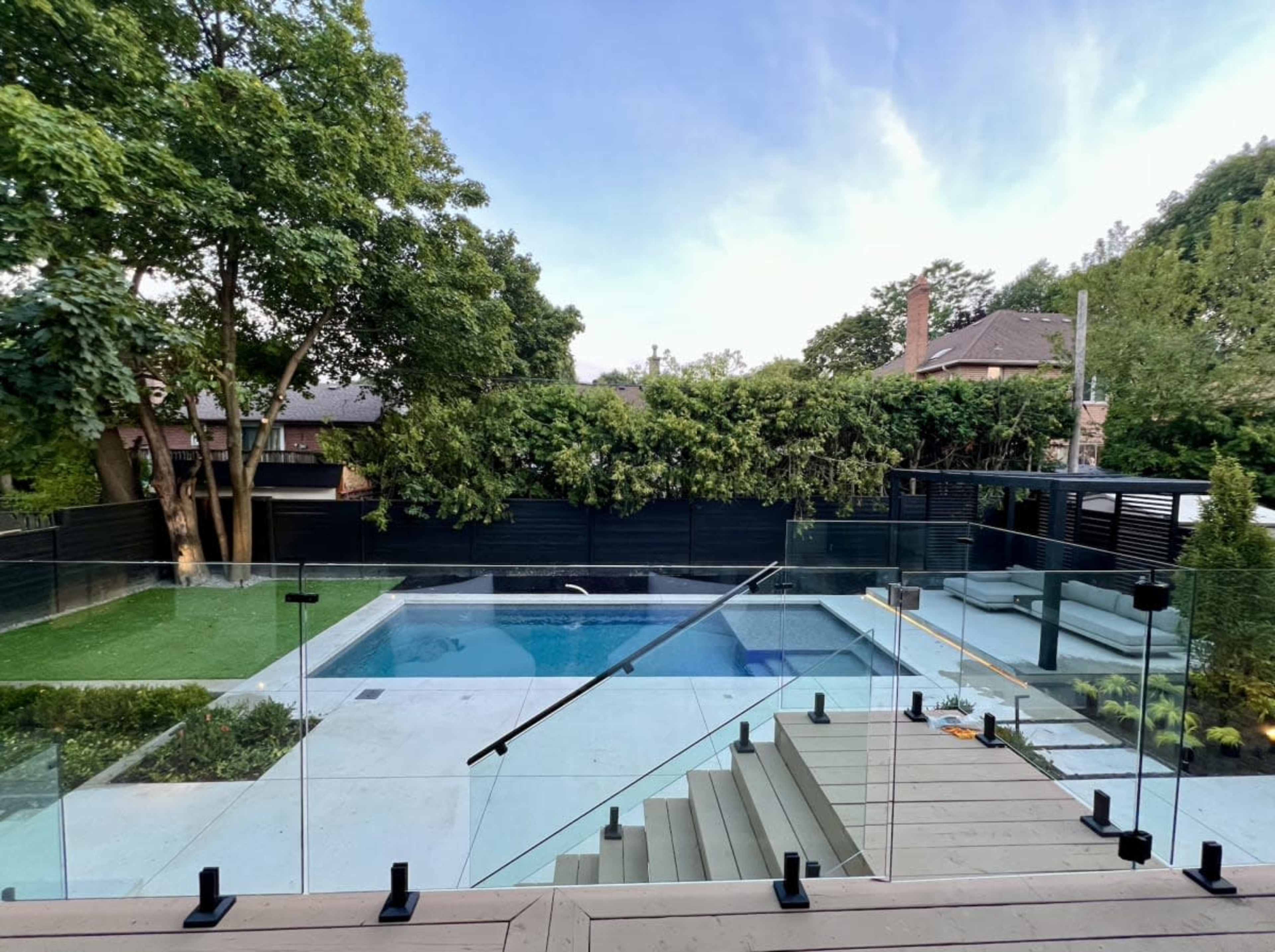 The image shows a backyard with a swimming pool bordered by glass railings, surrounded by greenery and landscaped areas.