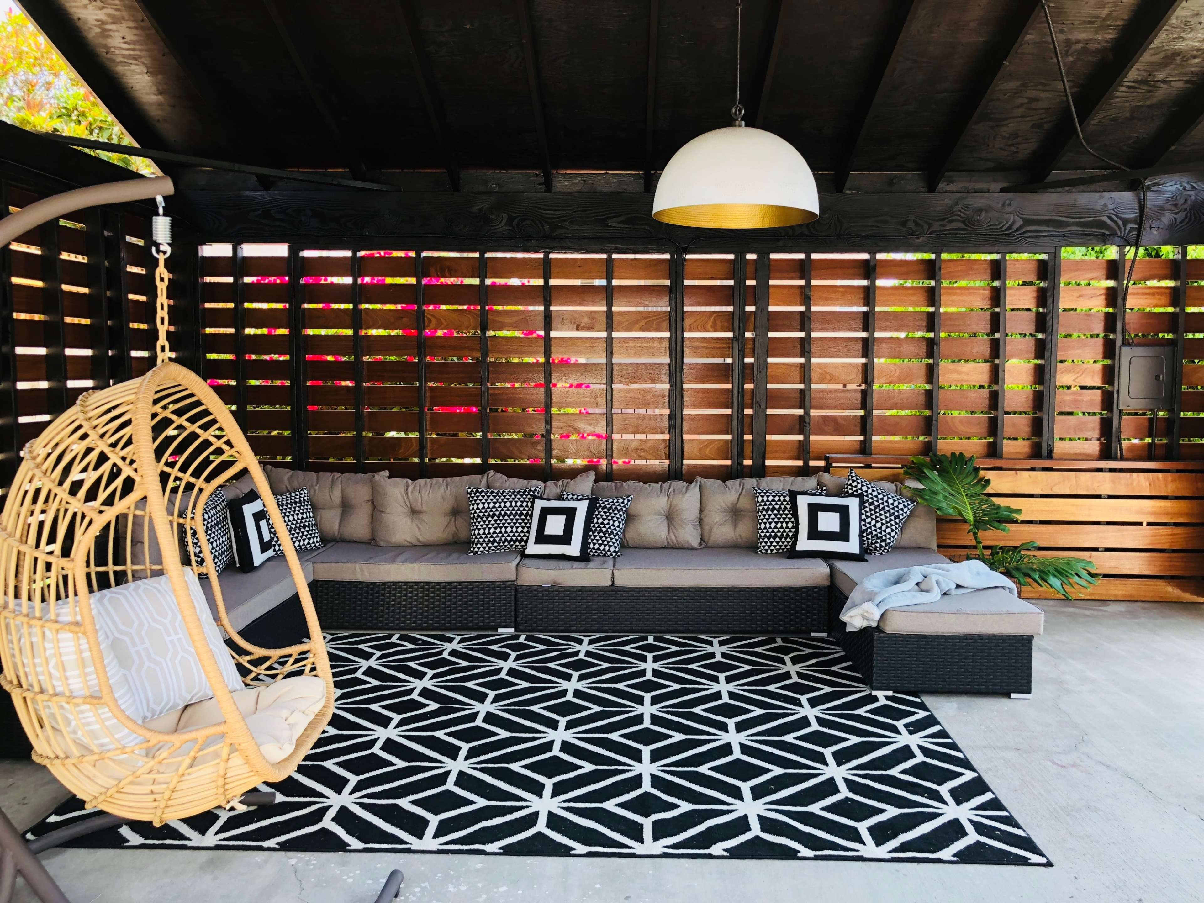 A shaded outdoor seating area with a sectional couch, decorative cushions, a hanging chair, and a geometric-patterned rug.