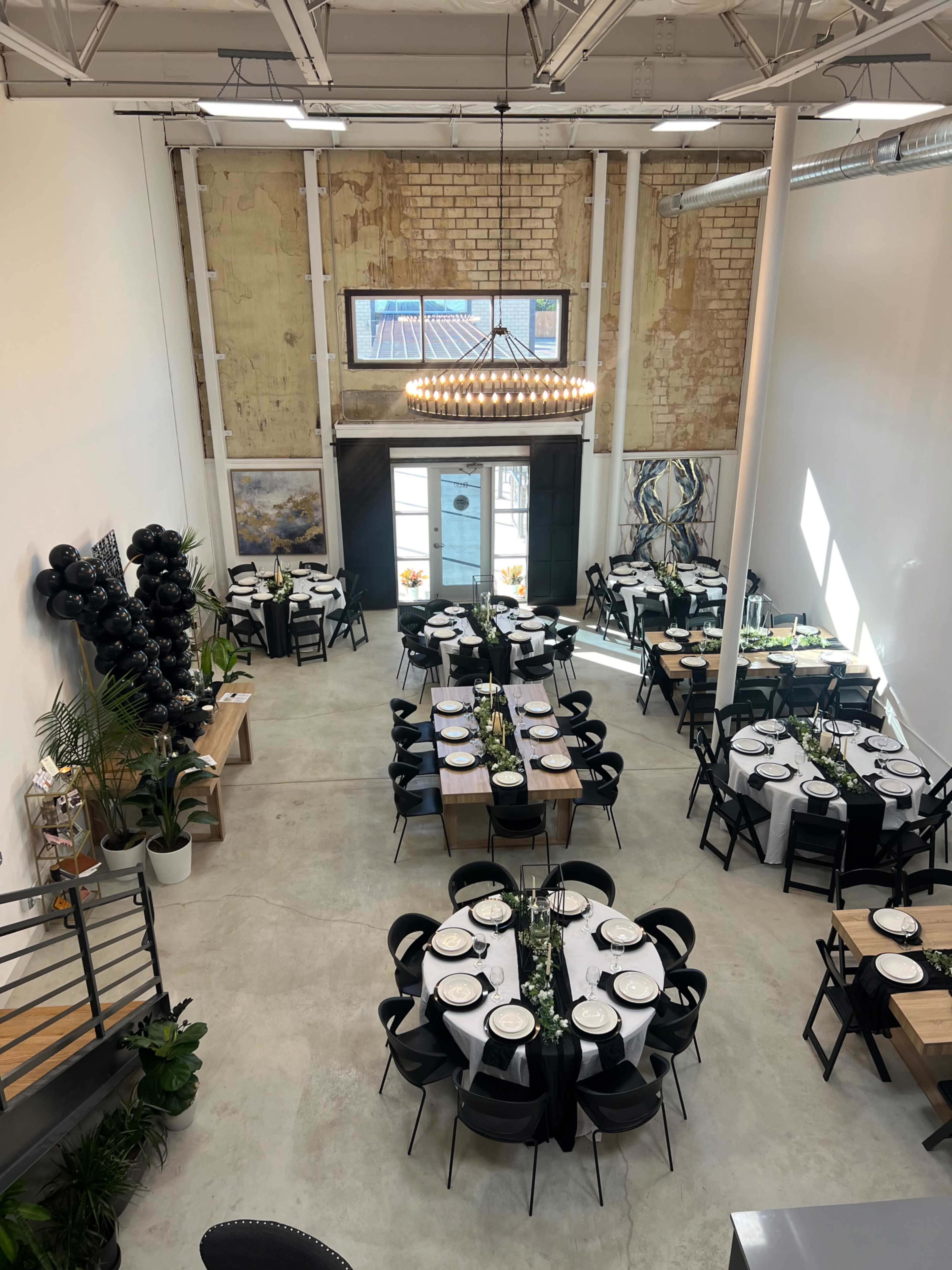 A spacious dining area with two round tables and several rectangular tables set for a formal event, featuring black chairs and a mix of decorative plants.