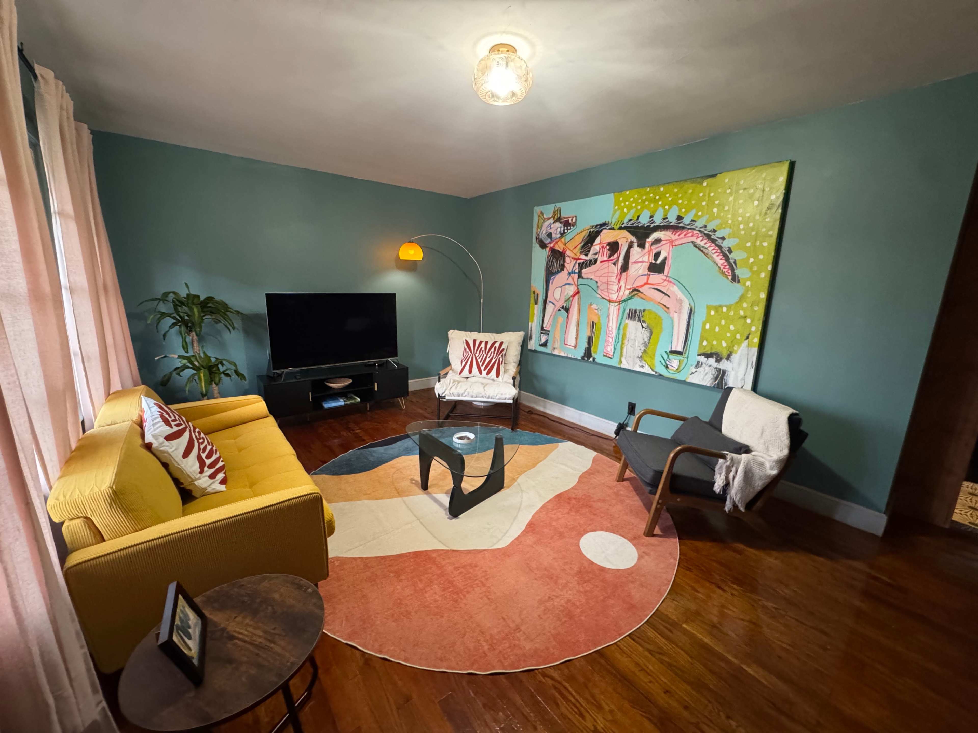 The image shows a modern living room with teal walls, a yellow sofa, a round area rug, and a large colorful artwork on one wall.