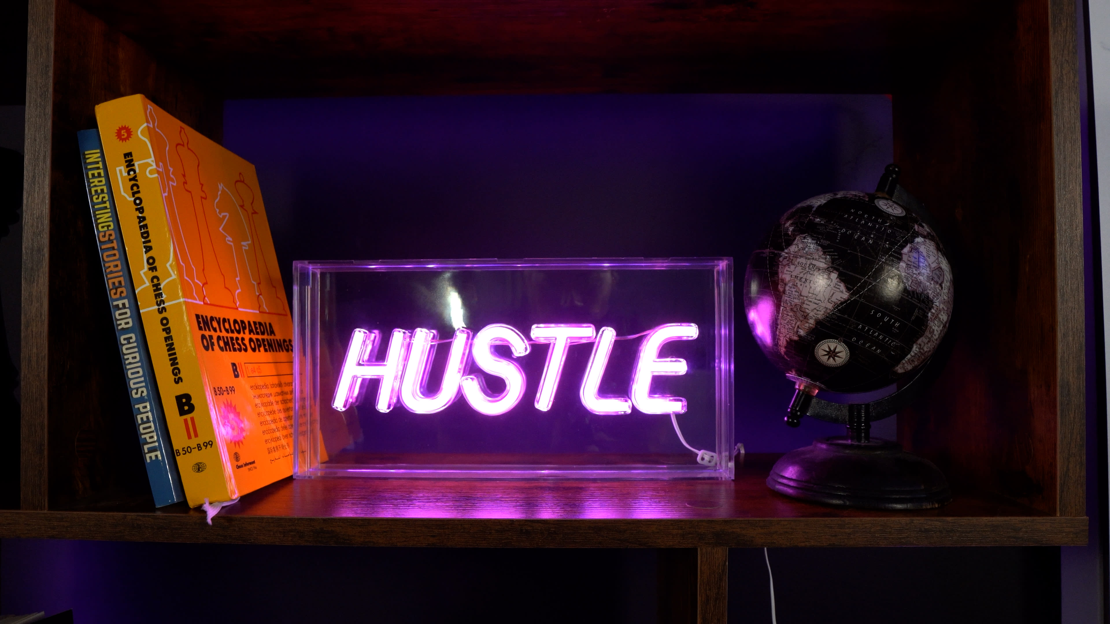 A neon sign that reads "HUSTLE" is illuminated on a shelf alongside books and a small globe.
