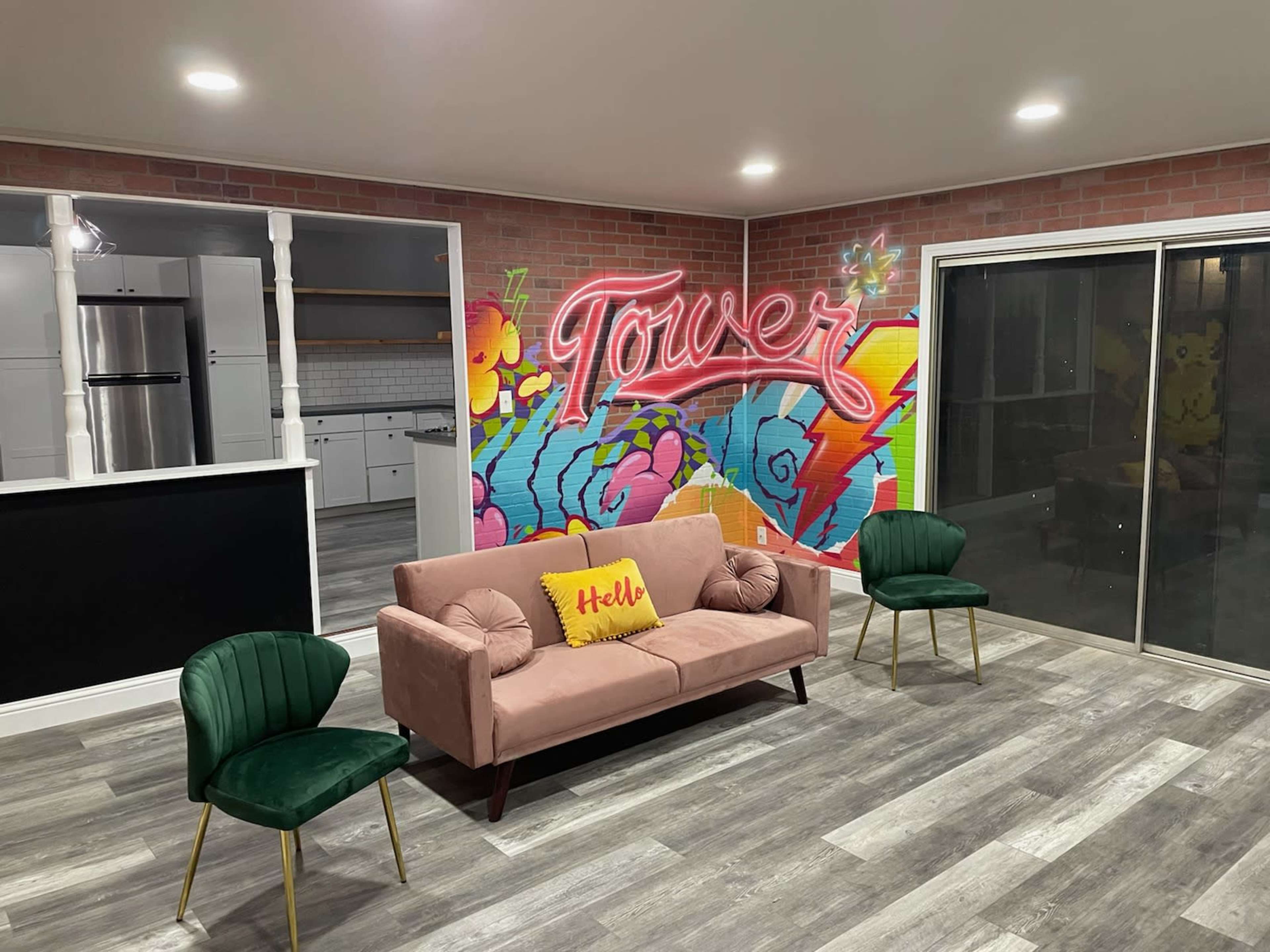 A modern living space features a pink sofa with a "Hello" pillow, two green chairs, and a colorful mural reading "Tower" on the wall.