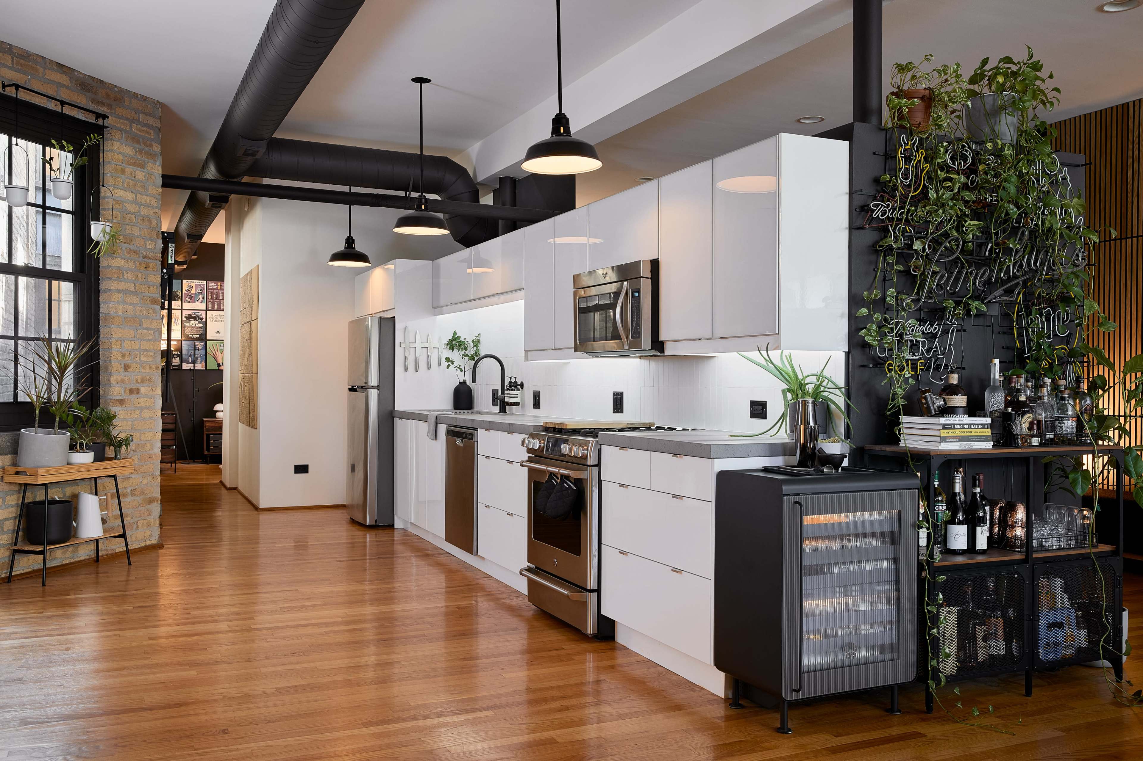The image shows a modern kitchen featuring white cabinetry, stainless steel appliances, and a dark accent wall with plants and a bar area, all set against a backdrop of hardwood flooring.