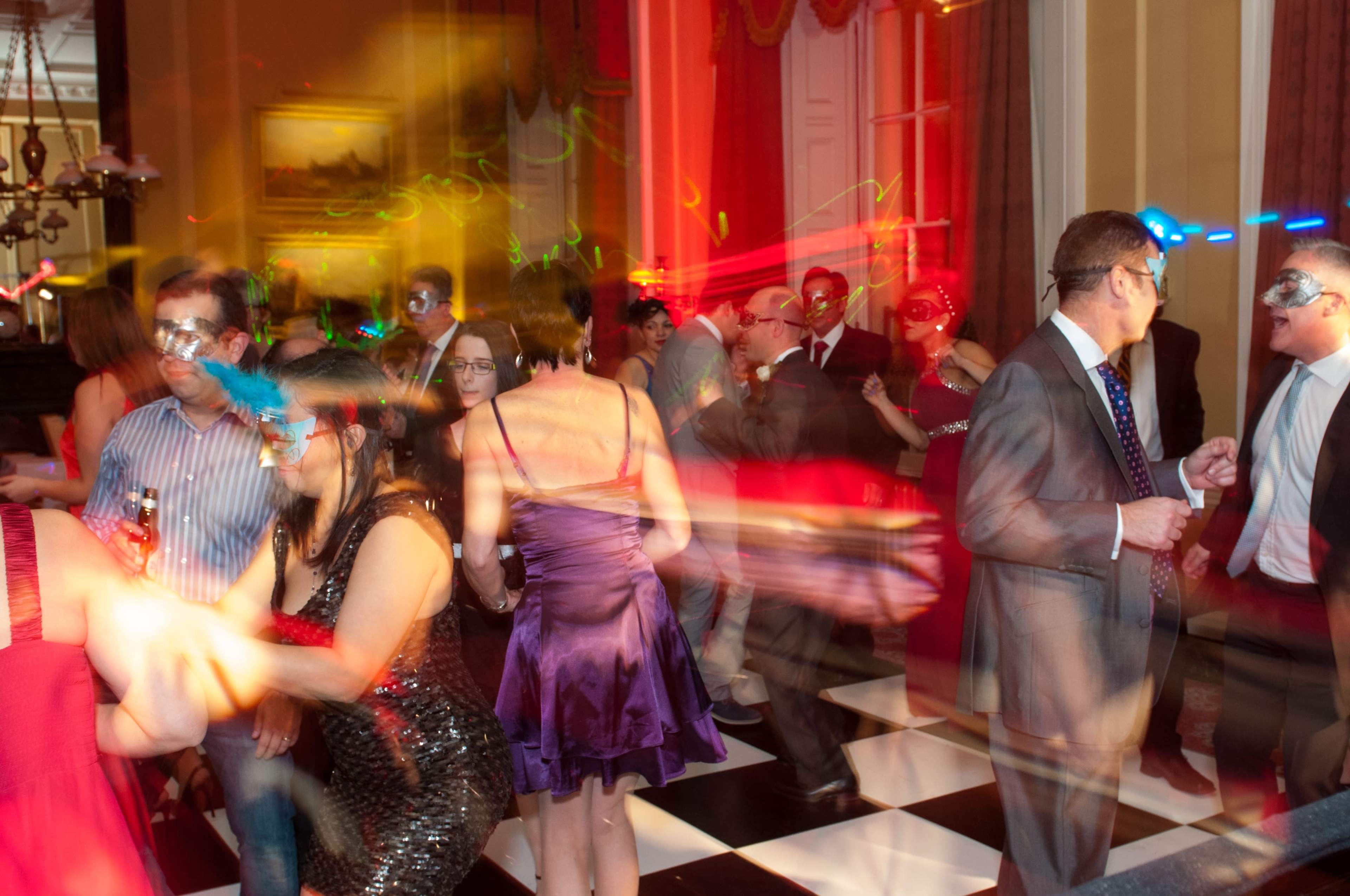 A lively dance party takes place in a elegantly decorated room with guests wearing masks and colorful lights creating streaks in the air.